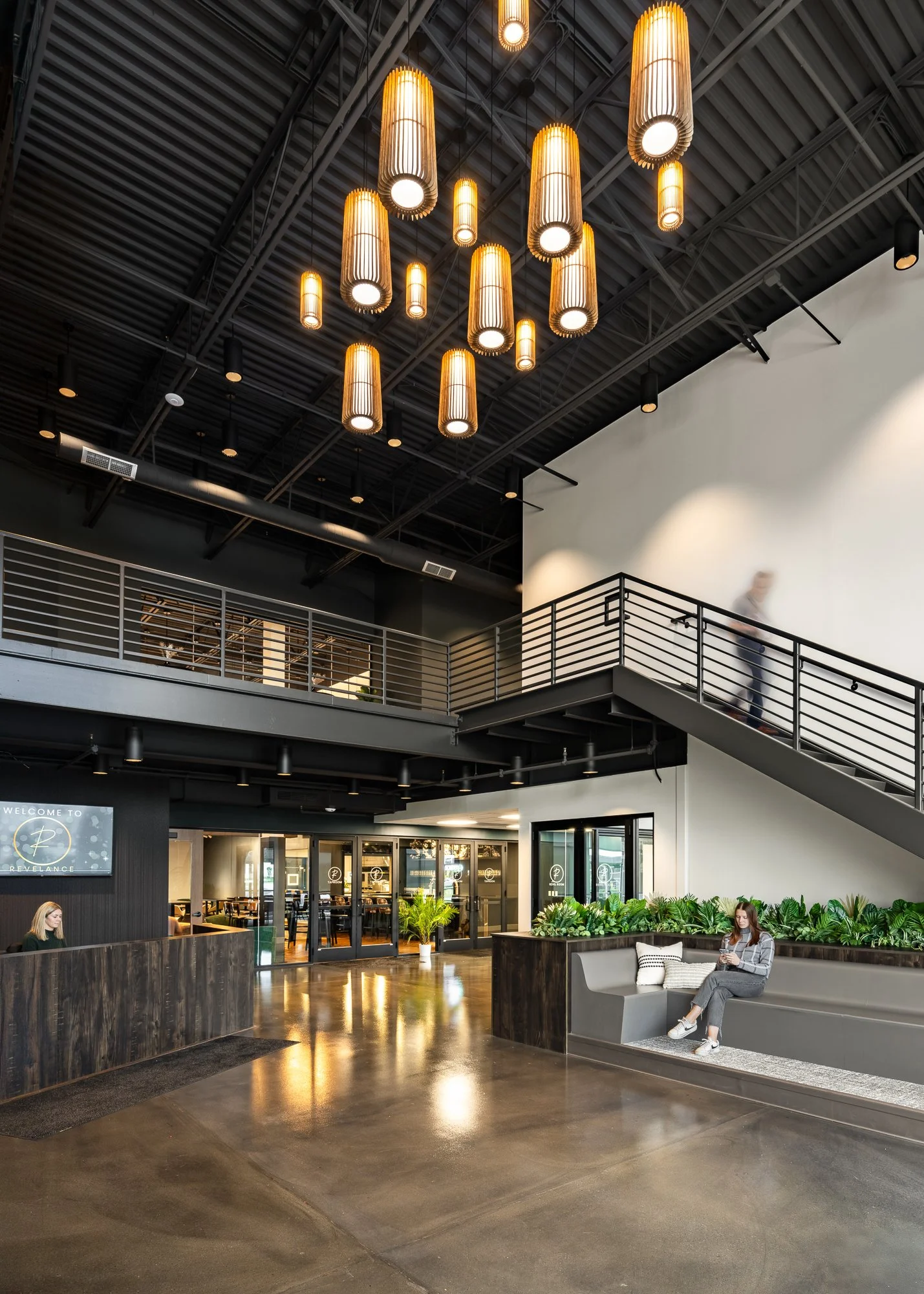Modern lobby with a high ceiling, hanging pendant lights, a reception desk, and a seating area with a woman using her phone.