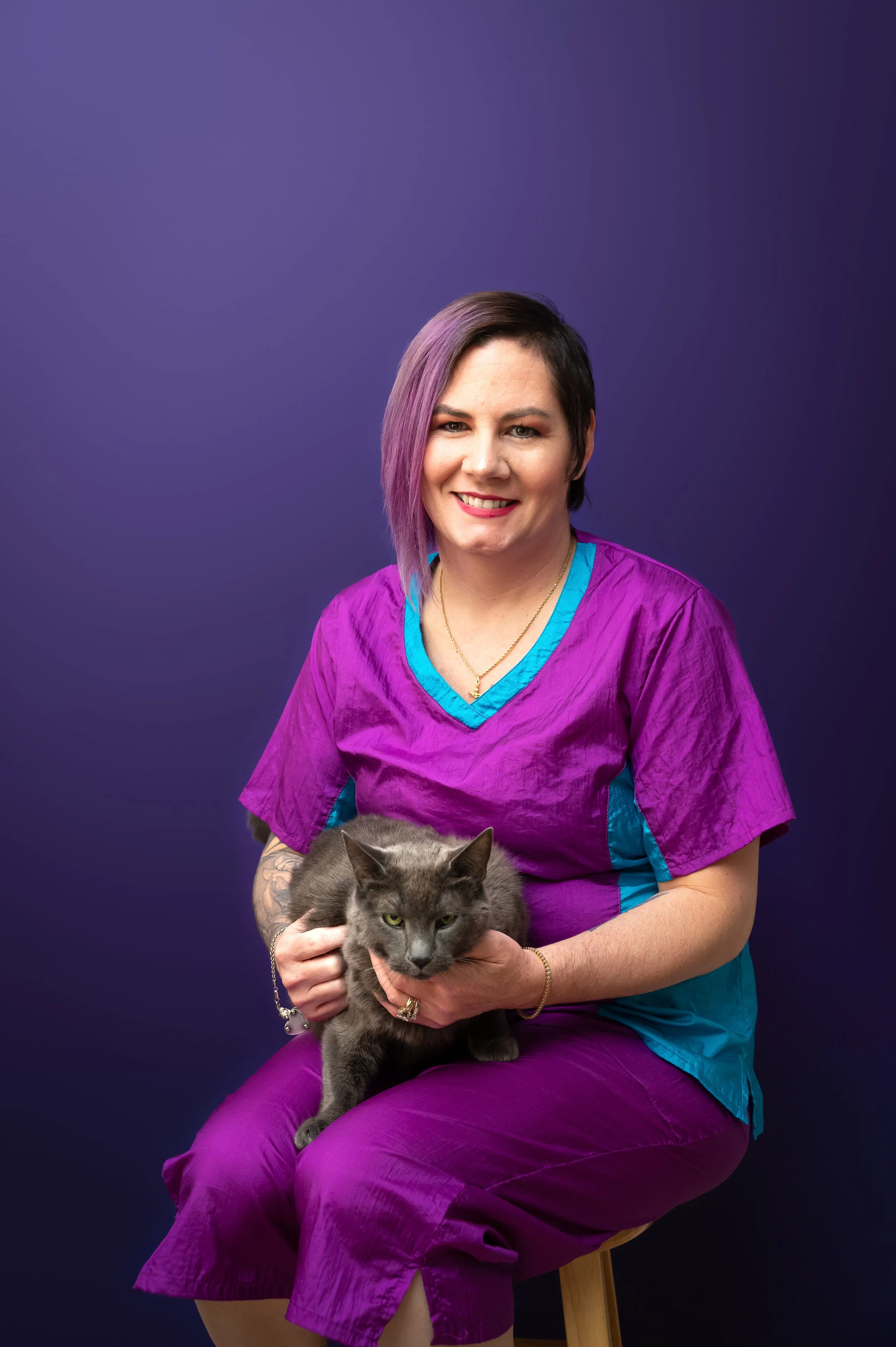 A woman with short, purple and black hair, wearing purple scrubs with blue accents, sitting on a wooden stool, holding a gray cat in her lap against a purple background.