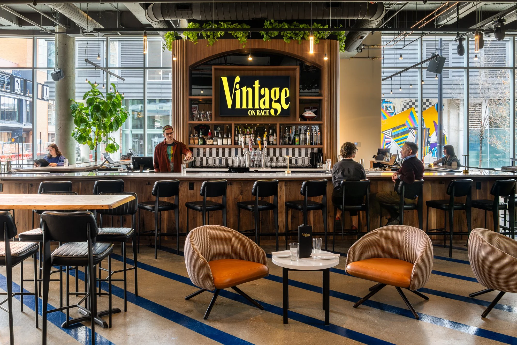 Interior of a modern bar named Vintage on Race, with a curved wooden bar counter, bar stools, and several patrons. A large screen displays the bar's name, surrounded by shelves with bottles and glasses. There are colorful geometric paintings on the w