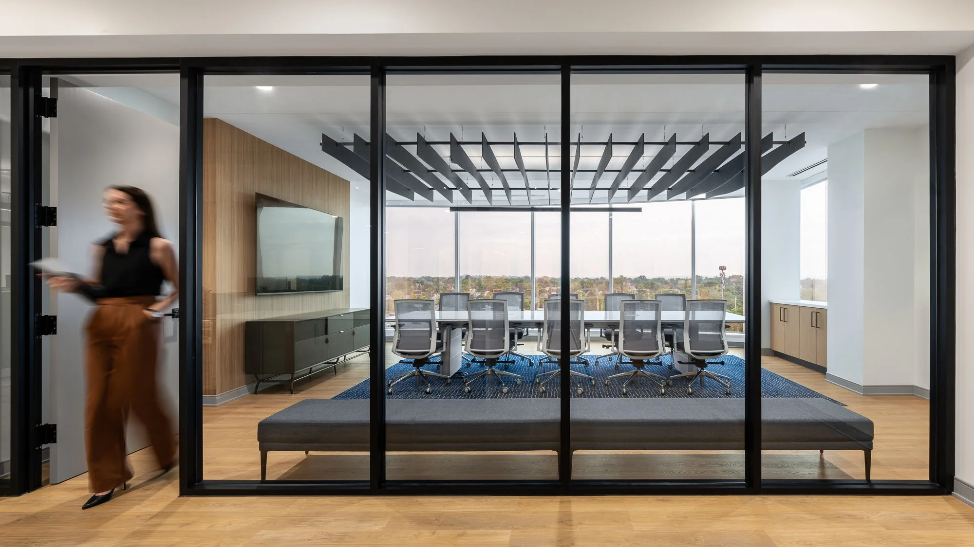 A modern conference room with glass walls, a long white table, and several office chairs. The room has large windows with a city view and a wood-paneled wall with a mounted TV.