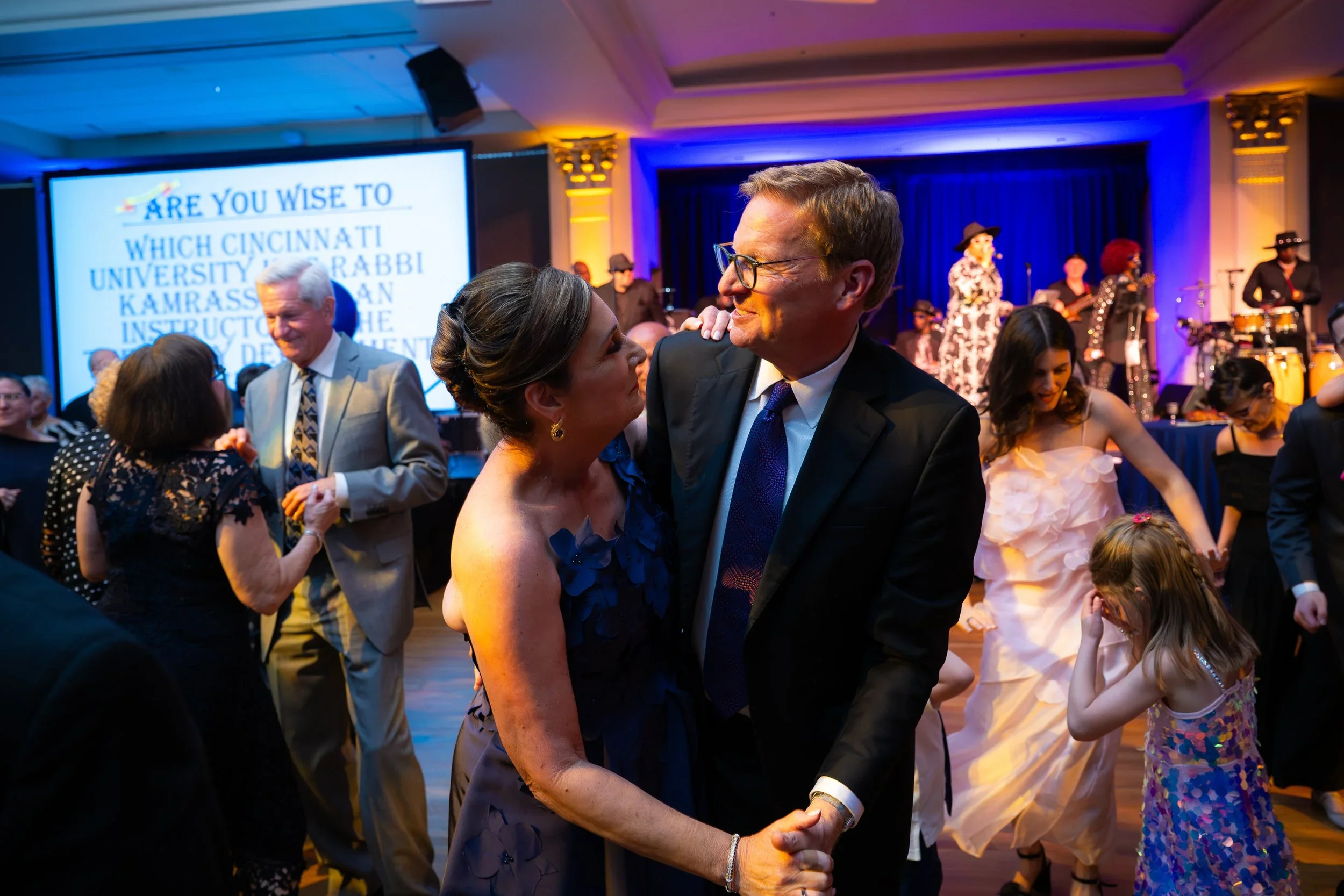 People dancing and socializing at a formal event with a band performing on stage and a large screen in the background.