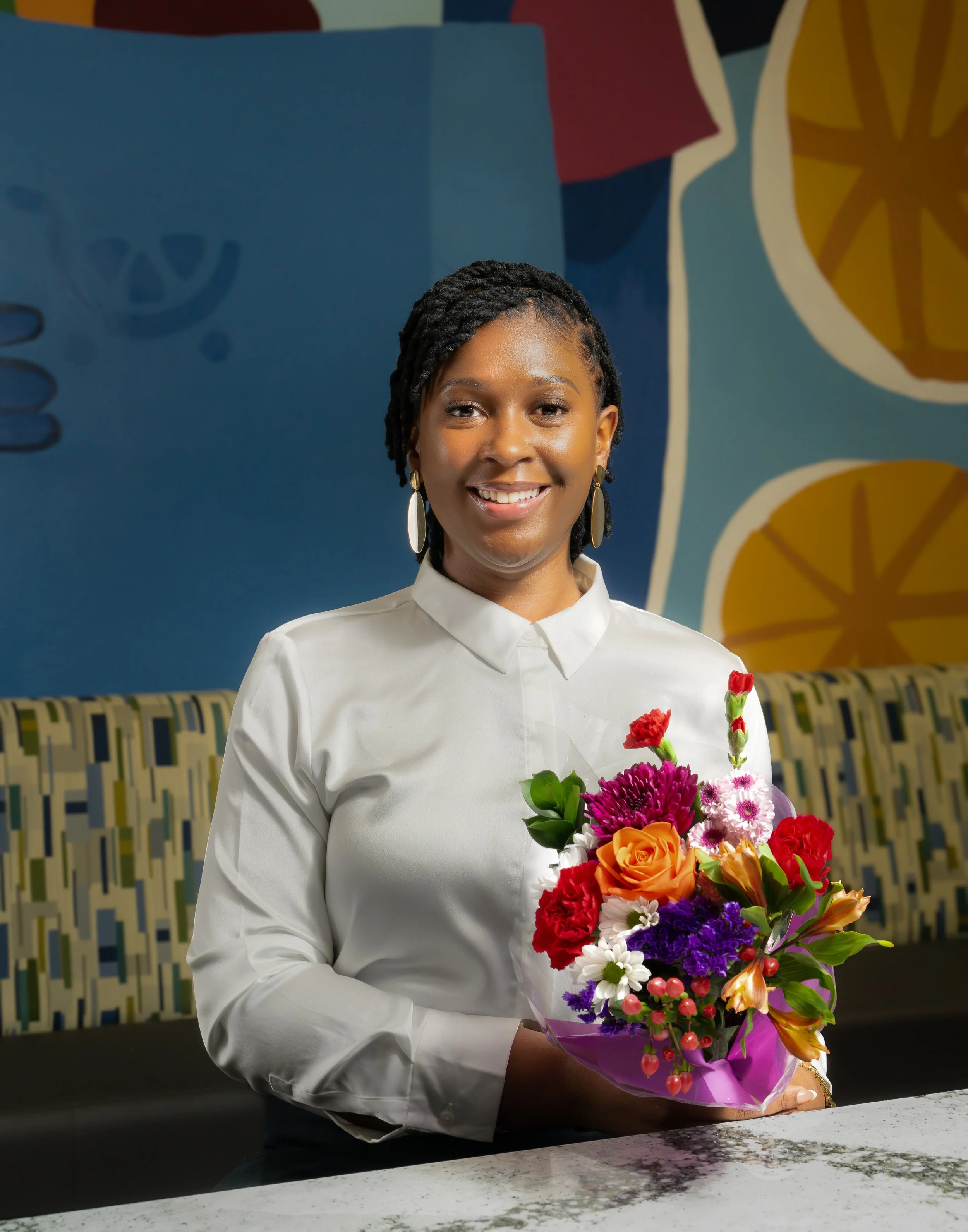 A young woman with short braided hair and earrings holding a colorful bouquet of flowers, standing in front of a vibrant mural background.