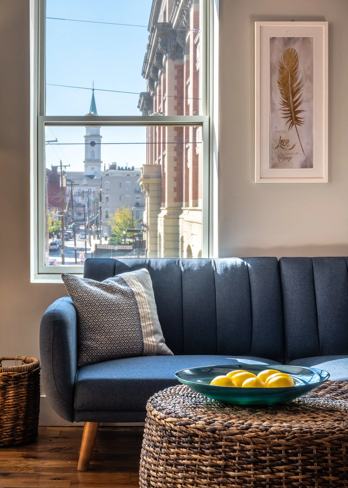 A living room with a dark blue sofa, a patterned pillow, a woven rattan ottoman with a green dish holding lemons, a wicker basket on the left, a window showing a street view with buildings and a church steeple, and a framed wall art with a feather an