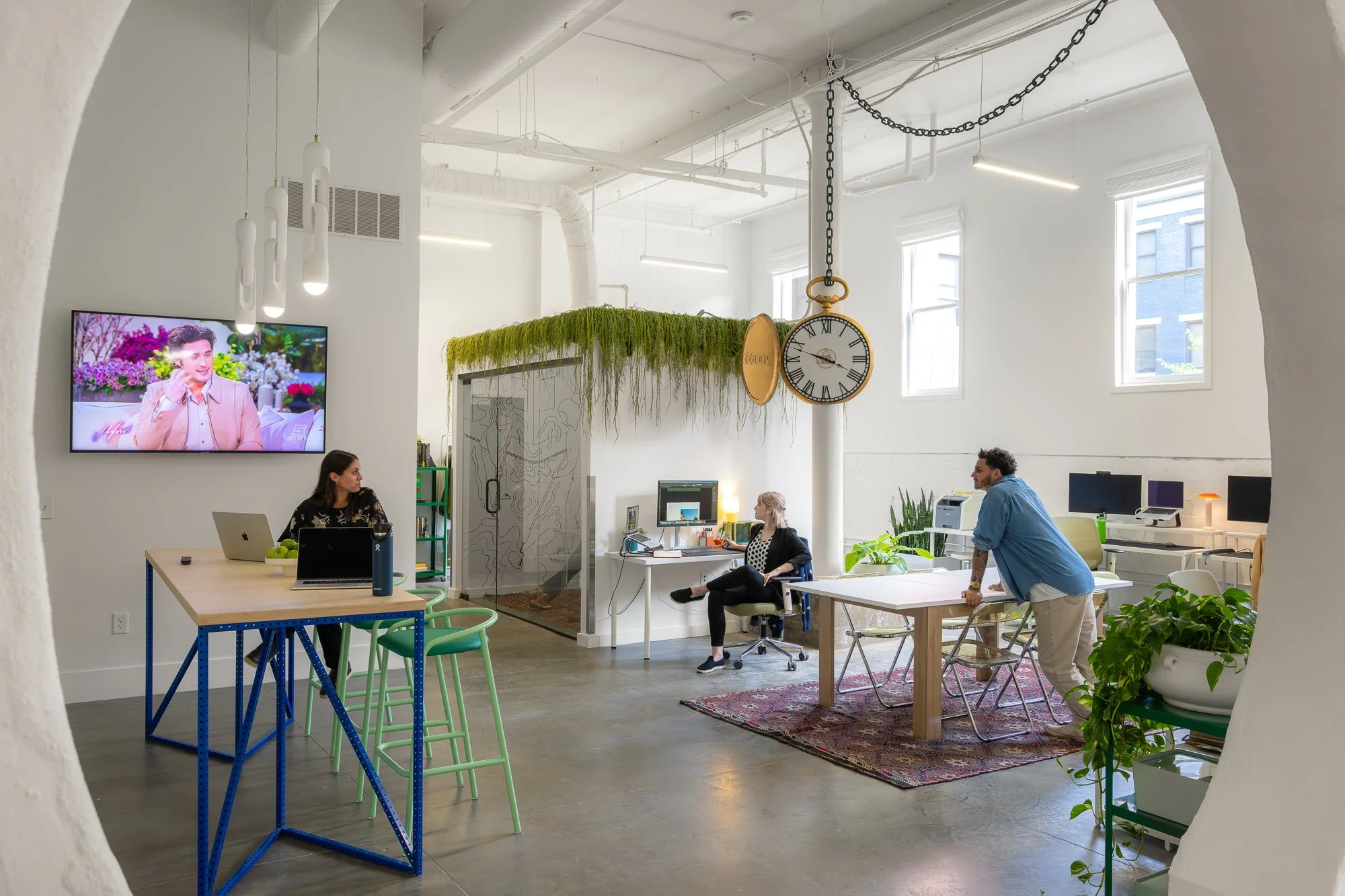 Open-plan coworking space with three people working at various desks, large windows, indoor plants, a mounted TV showing a person, clock hanging from the ceiling, and modern lighting.