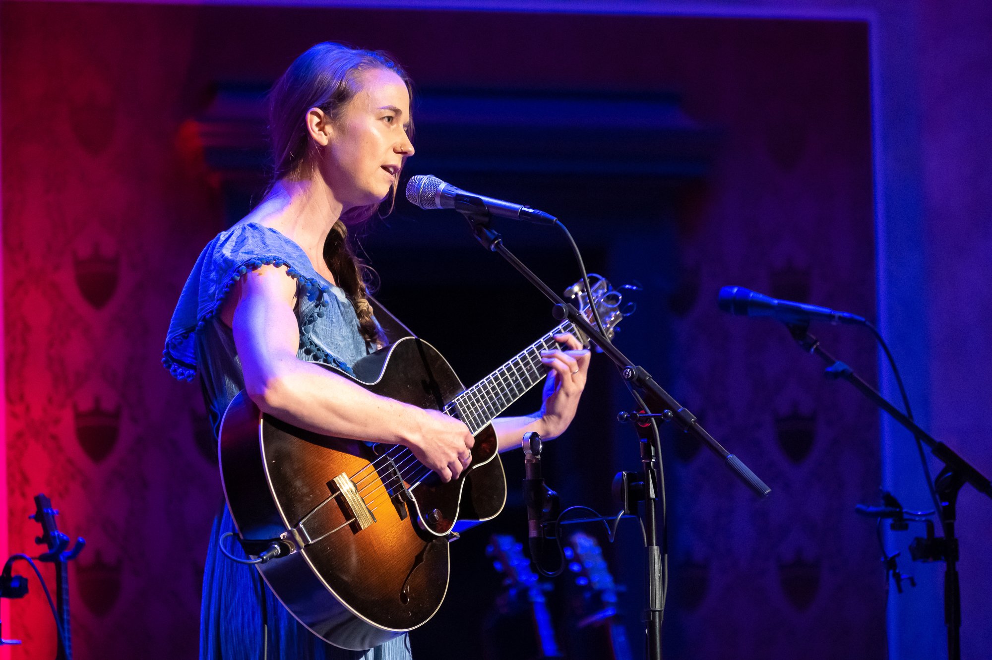 Woman performing on stage with a guitar and microphones, colorful backdrop with shield-shaped motifs.
