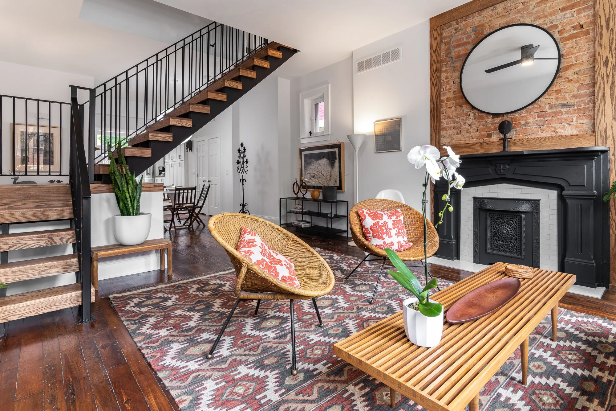 Living room with wooden floors, colorful patterned rug, two wicker chairs with orange and white cushions, a wooden slatted coffee table with a white orchid plant, black fireplace with a large round clock above, staircase with black railings and woode