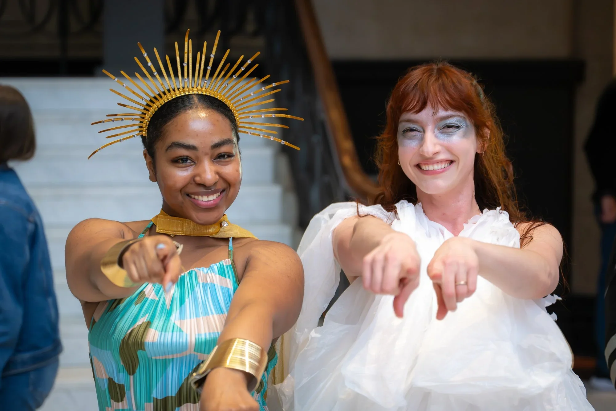 Two women smiling and pointing at the camera, one dressed in a colorful costume with a gold headpiece and the other in a white dress with silver makeup.