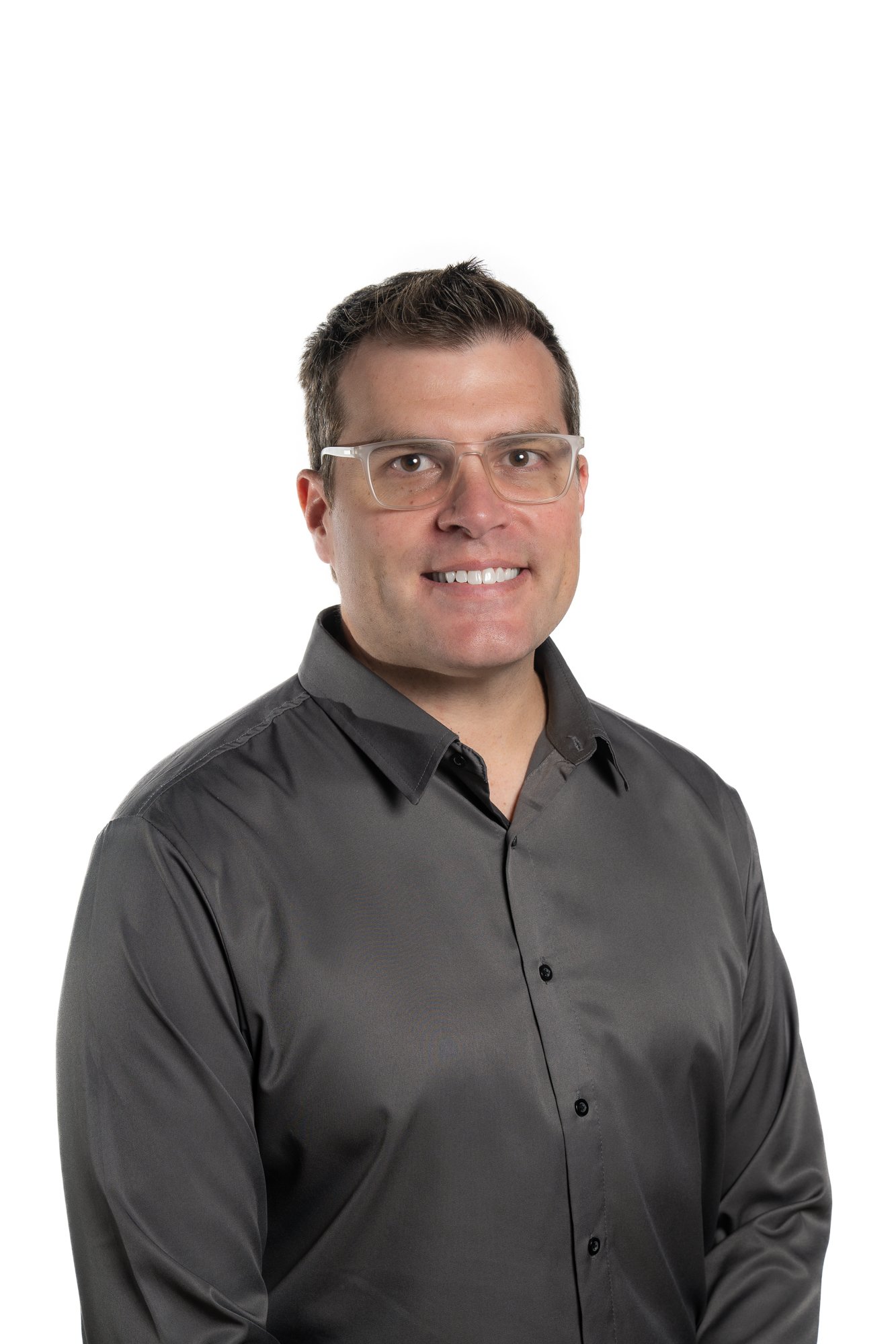 A man with short dark hair, glasses, and a friendly smile, wearing a dark gray button-up shirt, standing against a white background.