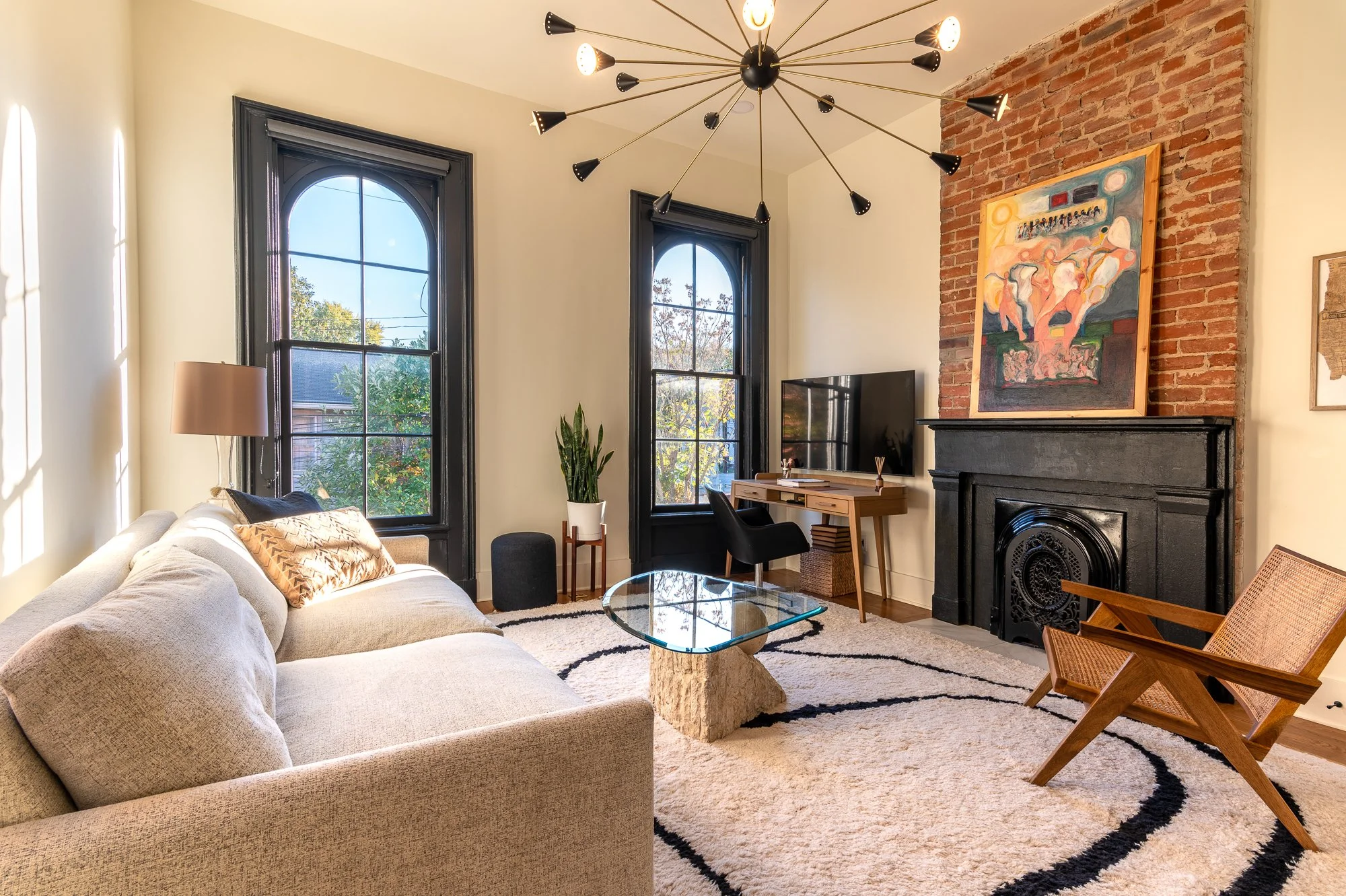 Modern living room with a beige sofa, glass coffee table on a white and black rug, brick fireplace, black framed windows, and contemporary artwork.