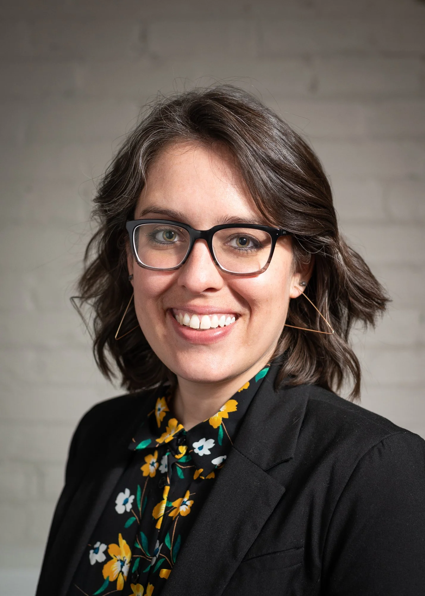 A woman with short brown hair, glasses, and earrings smiling in front of a neutral gray brick wall, wearing a black blazer and a floral blouse.