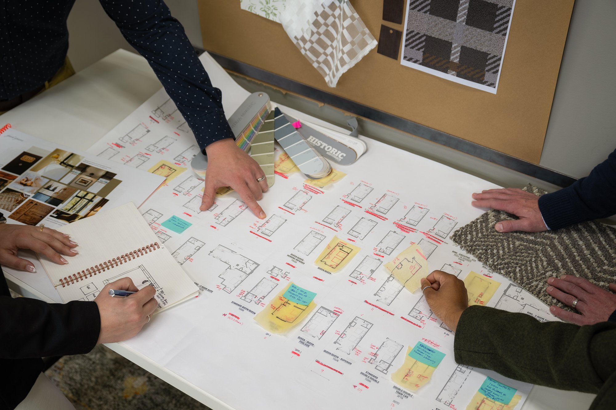 People working on architecture plans with floor plans, color samples, fabric swatches, and images of interior design ideas on a table.