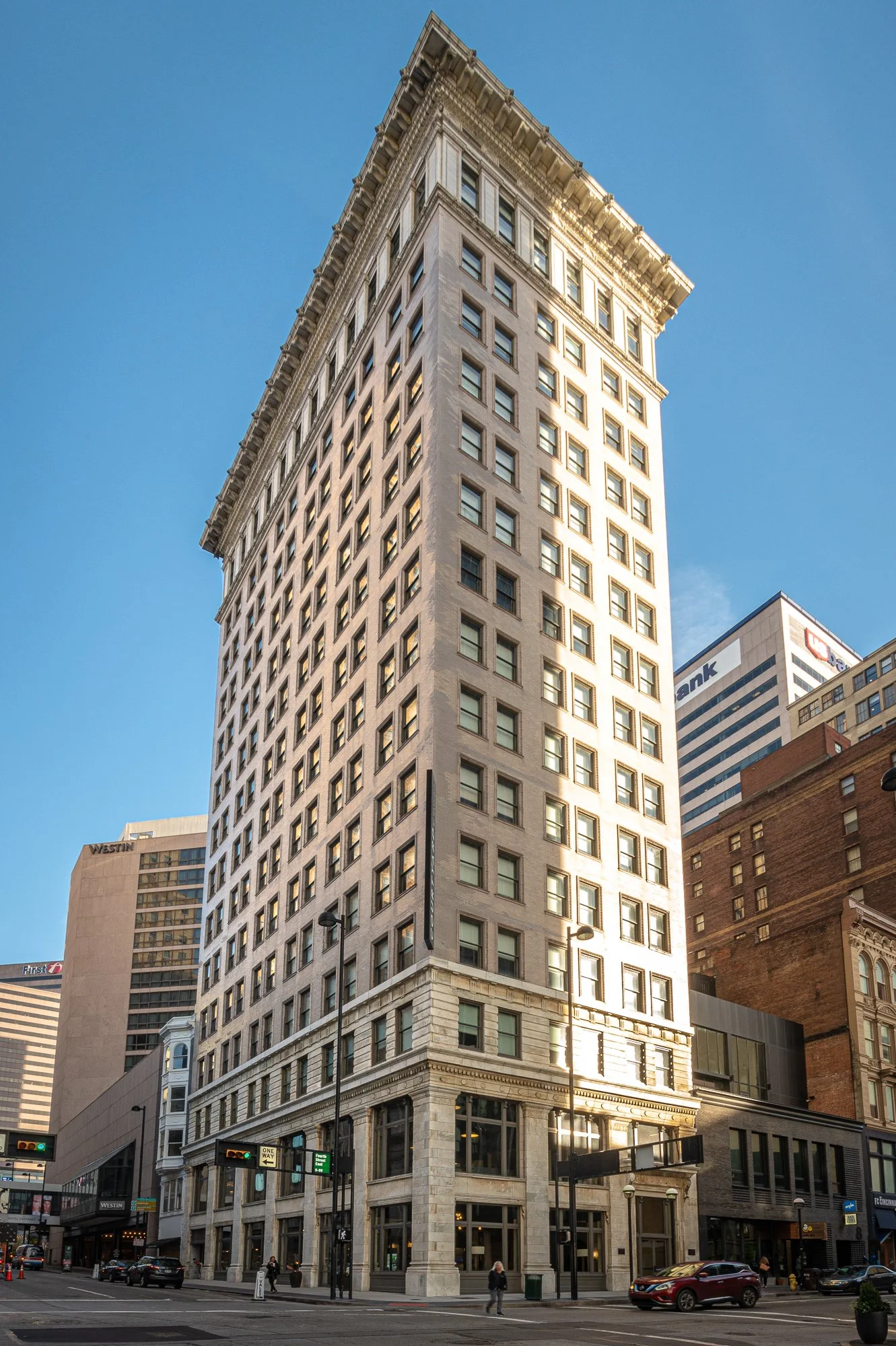 A tall, historic high-rise building with grid-like windows, located on a city street with cars and pedestrians, sunny weather, and other modern buildings in the background.