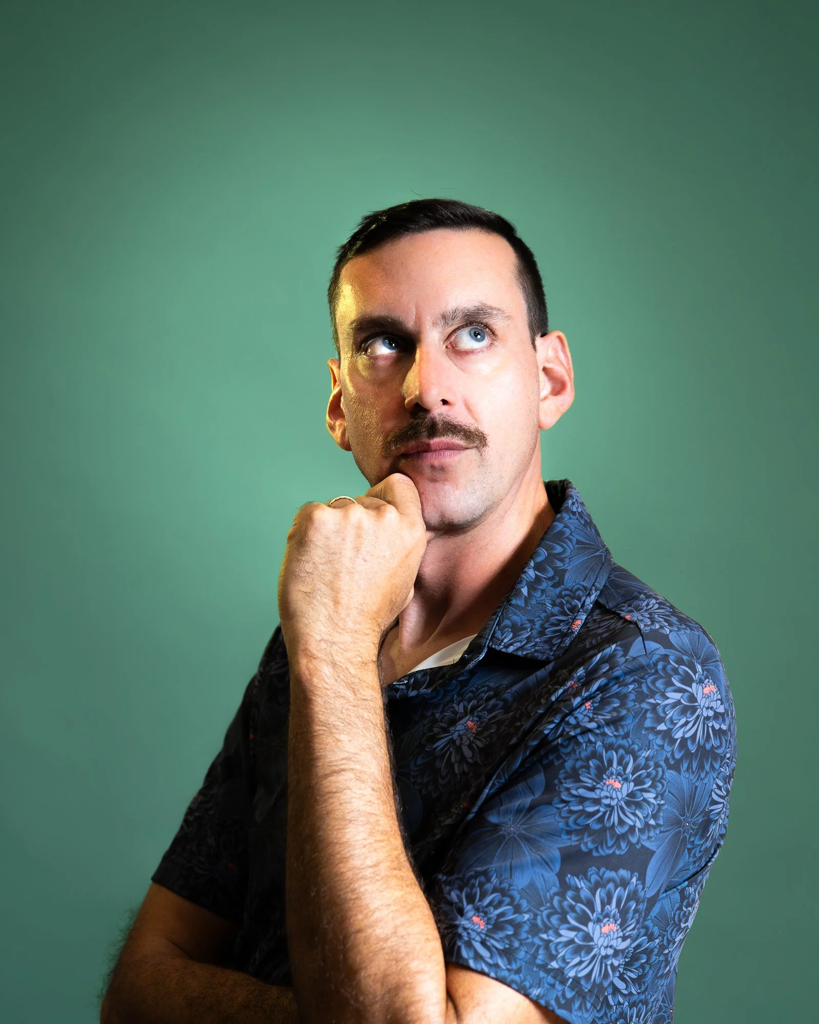 A man with dark hair, mustache, and light skin, wearing a blue floral shirt, posing against a green background with his hand on his chin, looking upwards.
