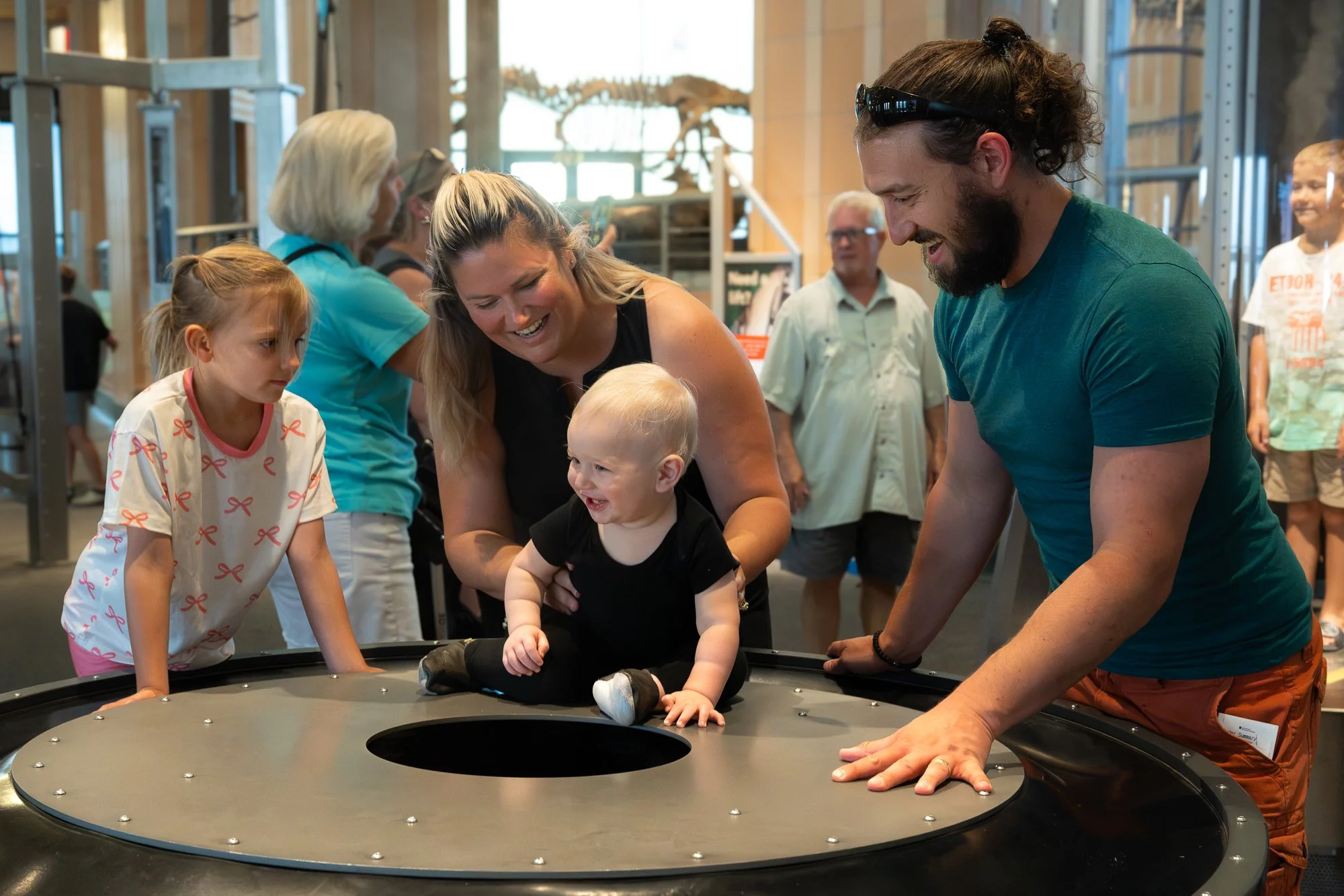 A group of people, including children and adults, gathered around a small trampoline or interactive exhibit in a museum, with a young child smiling as an adult woman and man support and encourage the child.
