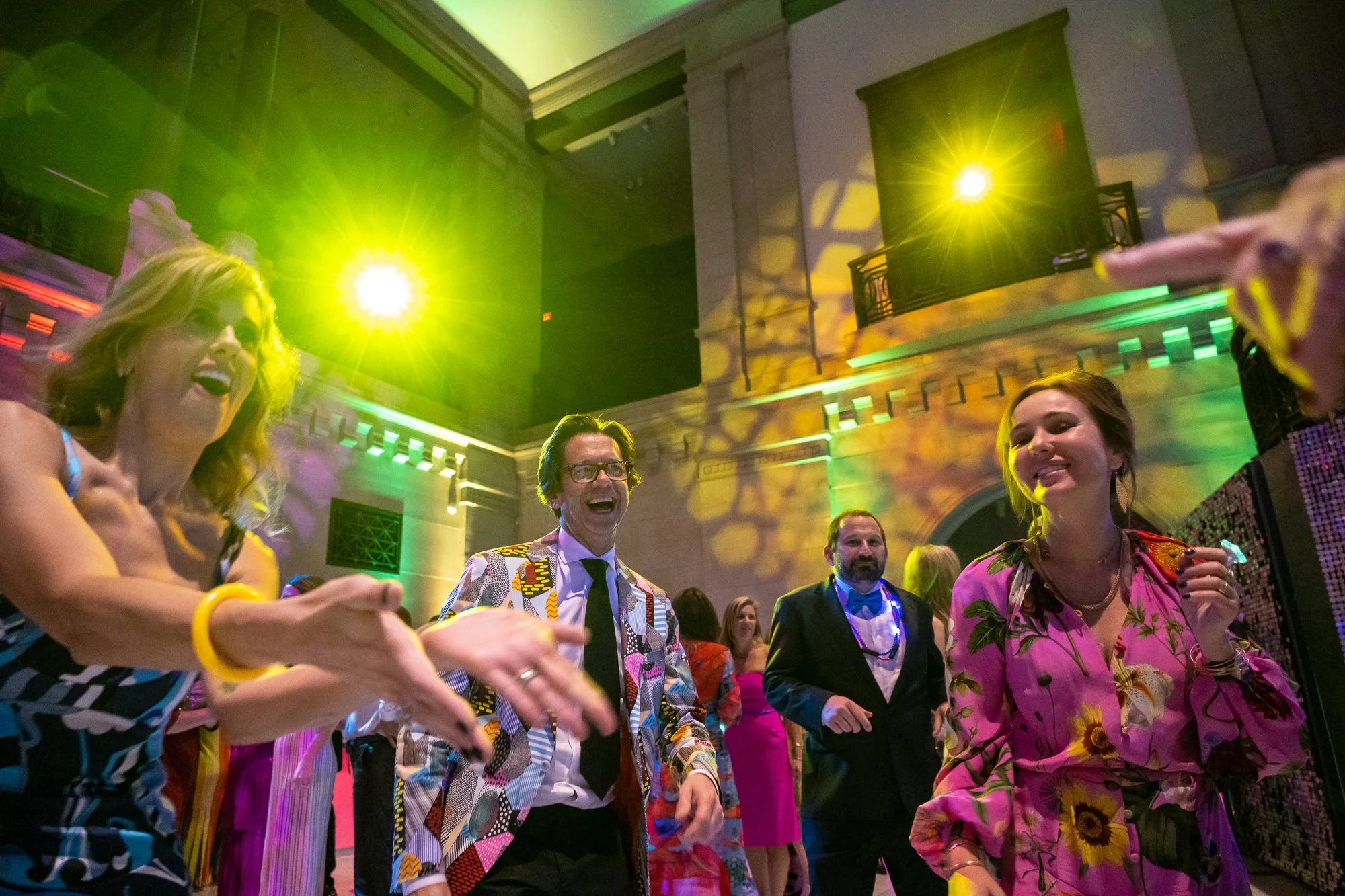 People dancing and laughing at a lively indoor party with colorful lighting and a high ceiling.