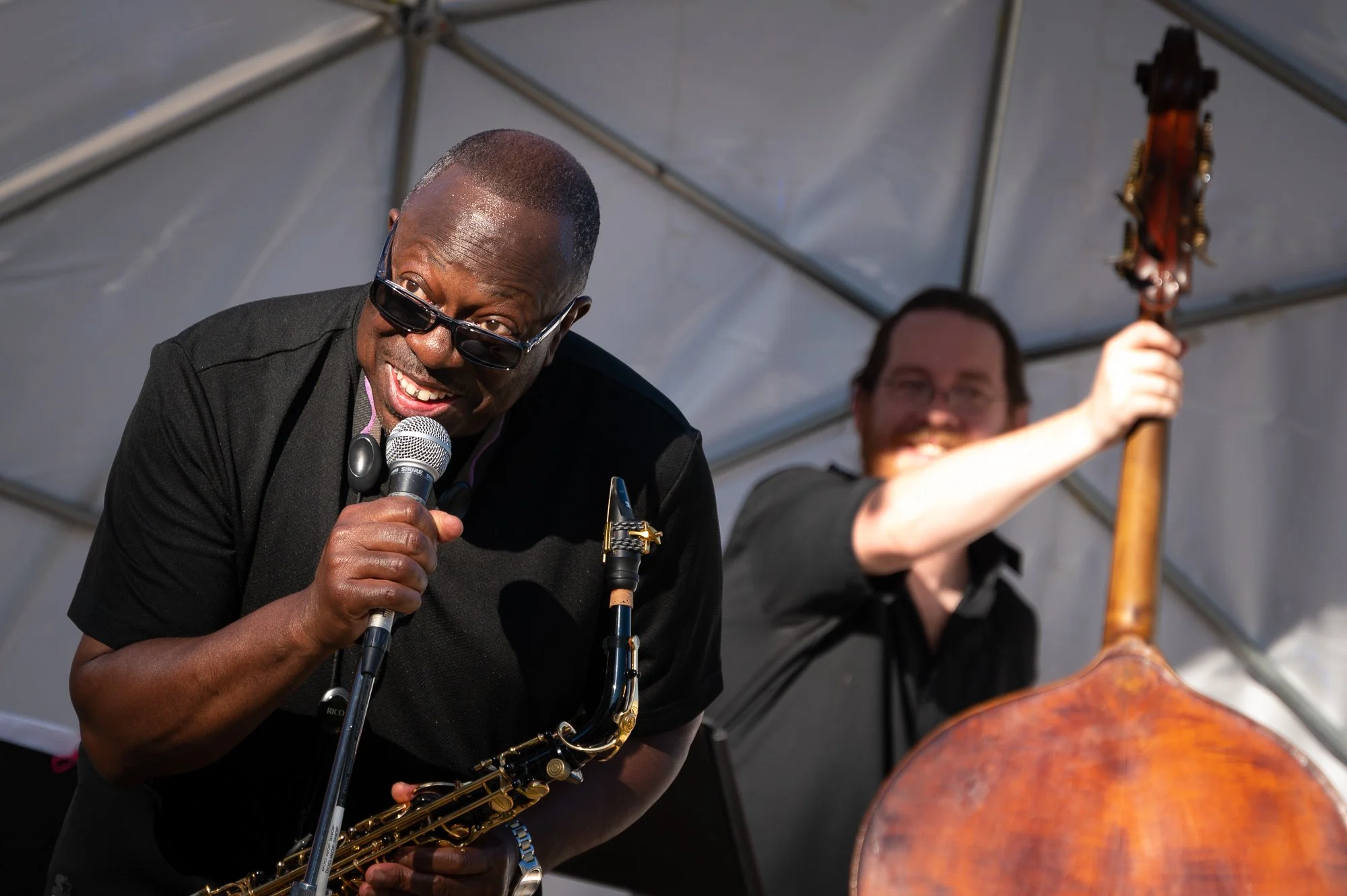 A man with glasses and a black shirt holding a microphone, playing a saxophone and smiling, with another man in the background holding a double bass and smiling, inside a metallic tent.