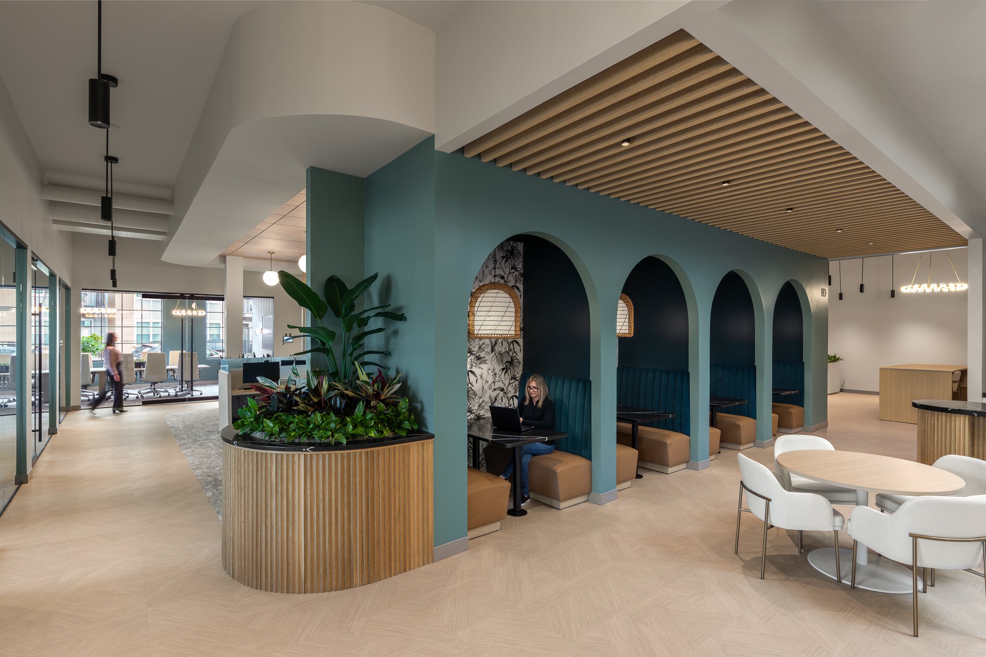 Modern office lounge area with booths, tables, and plants, featuring teal walls and wooden accents.