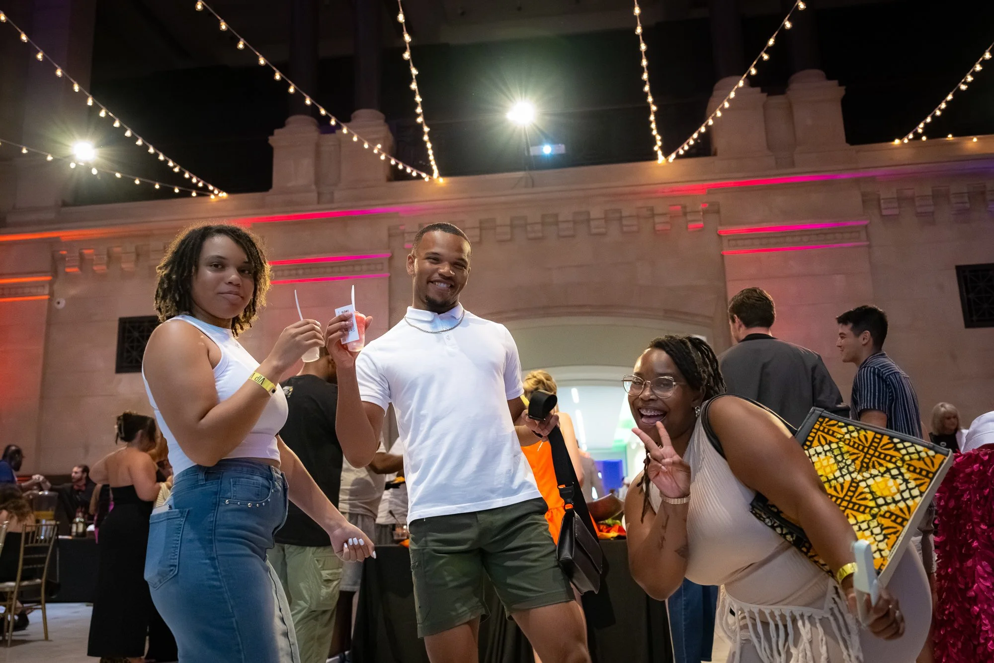 Three smiling people at a social event, standing and posing for the camera under string lights and a decorated archway, with other attendees in the background.