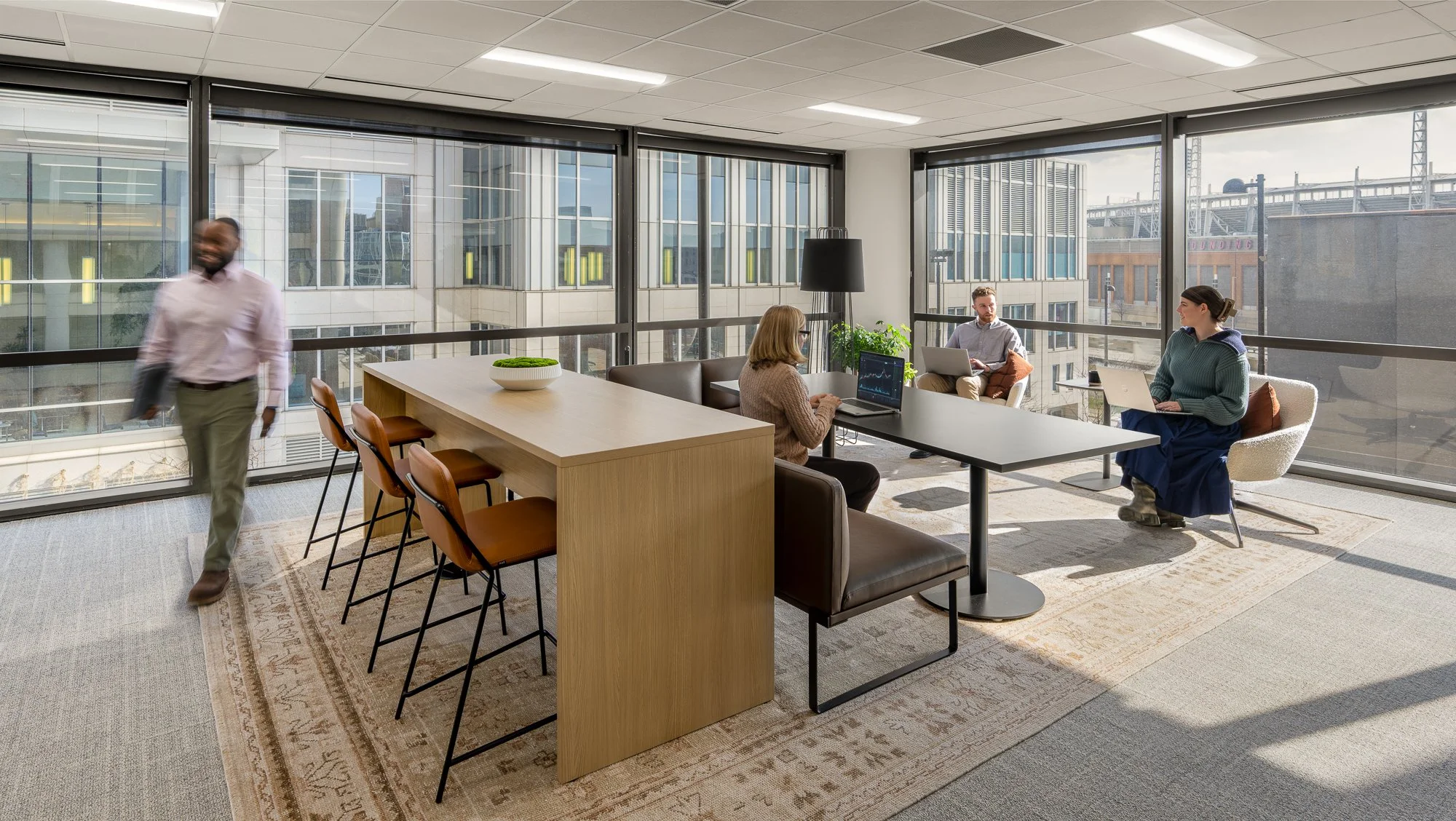 Office with large windows, four people working at a table, one walking, and various chairs and decor.