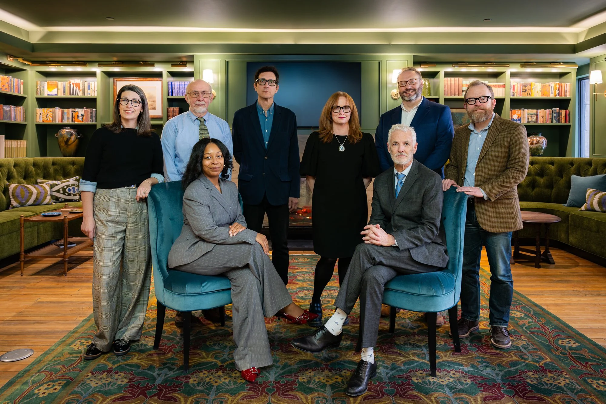 A group of ten professionally dressed people in a living room with a fireplace, bookshelves, and green walls.