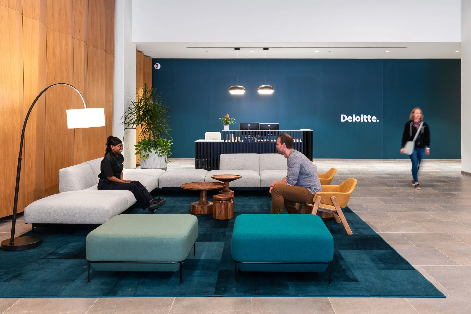Deloitte office lobby with seating area, two people talking, and a woman walking in the background near the Deloitte sign.