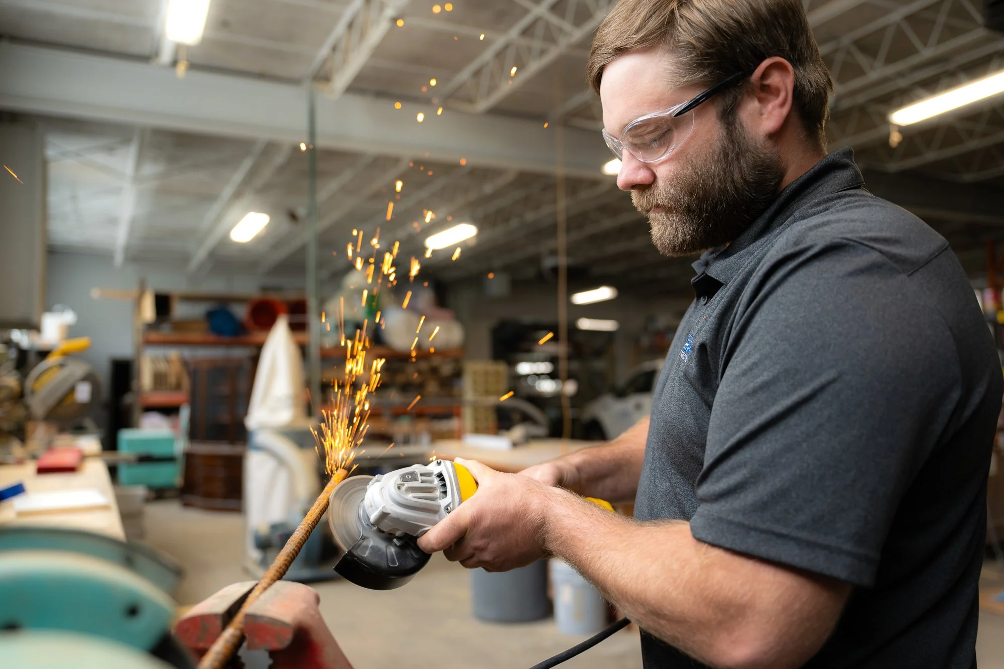 A man wearing safety glasses using an angle grinder to cut or grind a metal rod, with sparks flying as he works in a workshop or industrial setting.