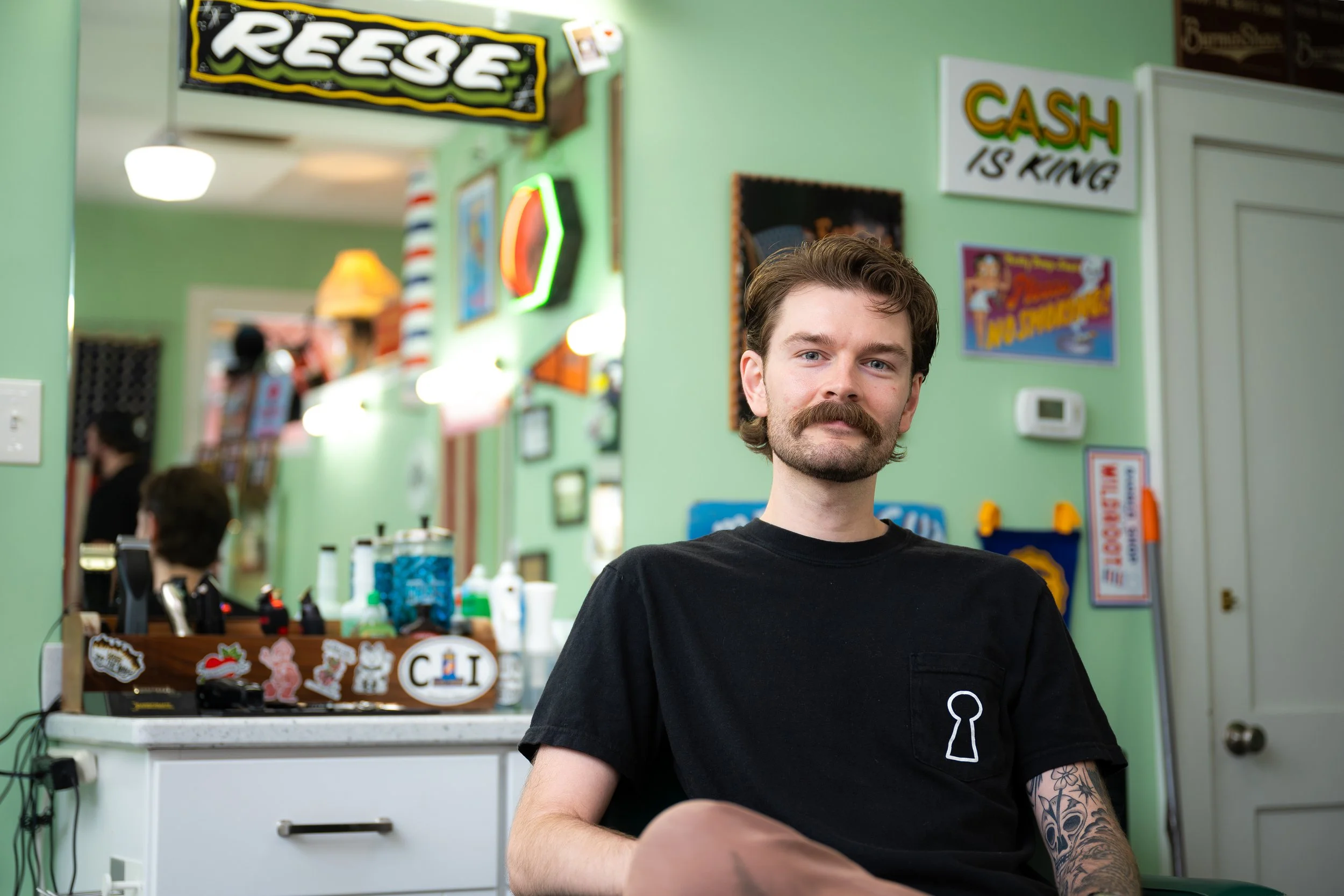 A young man with a mustache and tattooed arm sitting in a barbershop or hair salon, wearing a black t-shirt with a white keyhole logo, with colorful signs and decorations in the background.