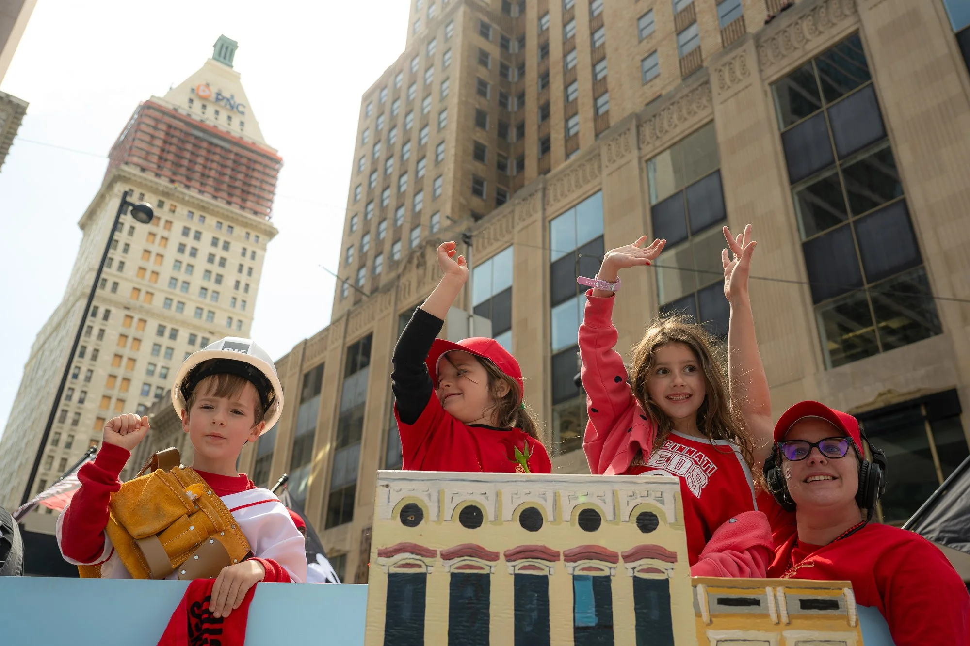Children and an adult on a parade float, wearing red shirts and hats, with a cityscape background of tall buildings and skyscrapers.
