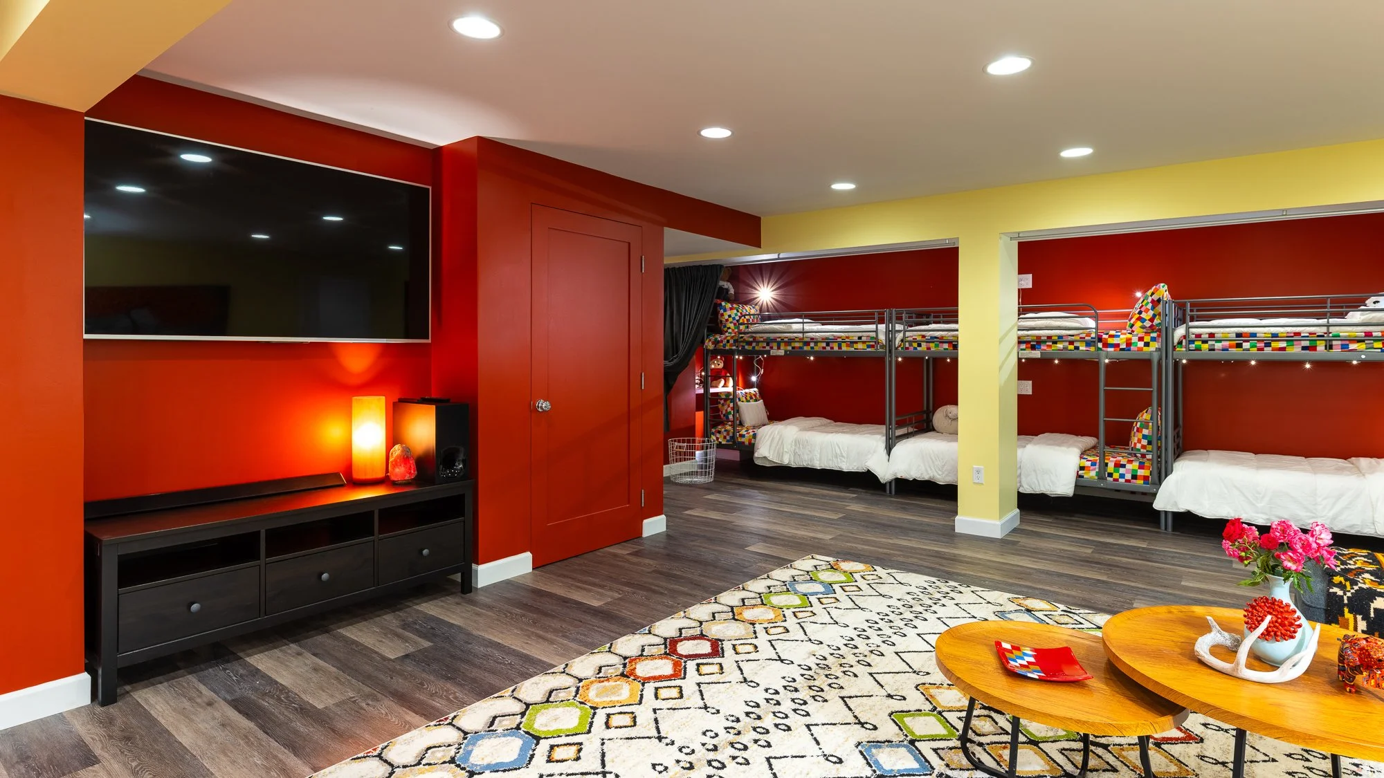 A colorful, multi-bedroom dormitory room with red and yellow walls featuring bunk beds with colorful patterned bedding and a large flat-screen TV.