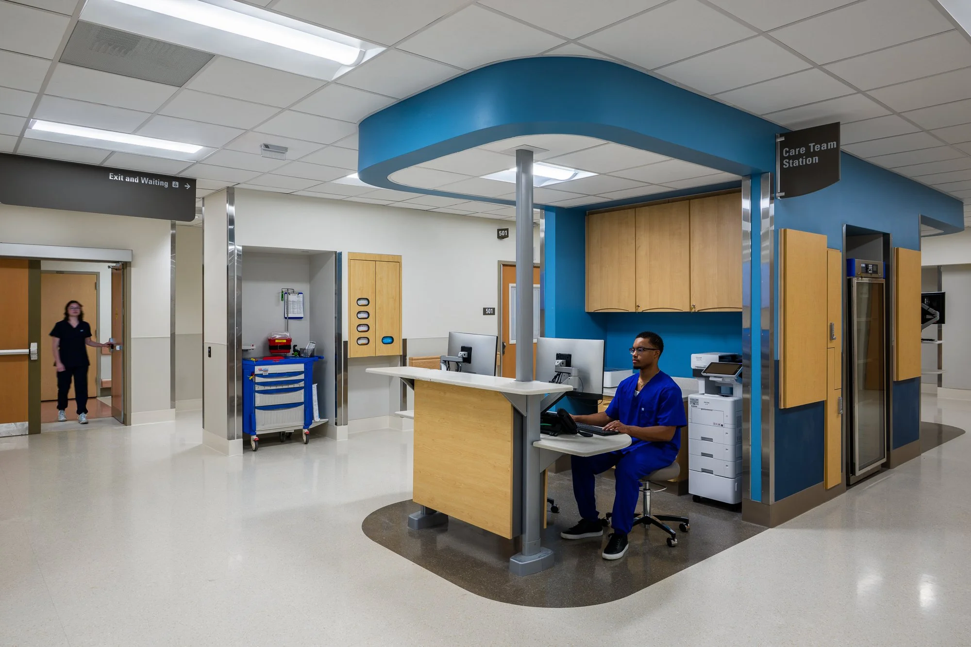 A hospital nurse sitting at a reception desk within a designated area labeled 'Care Team Station,' with medical equipment and cabinetry around, in a hospital corridor.