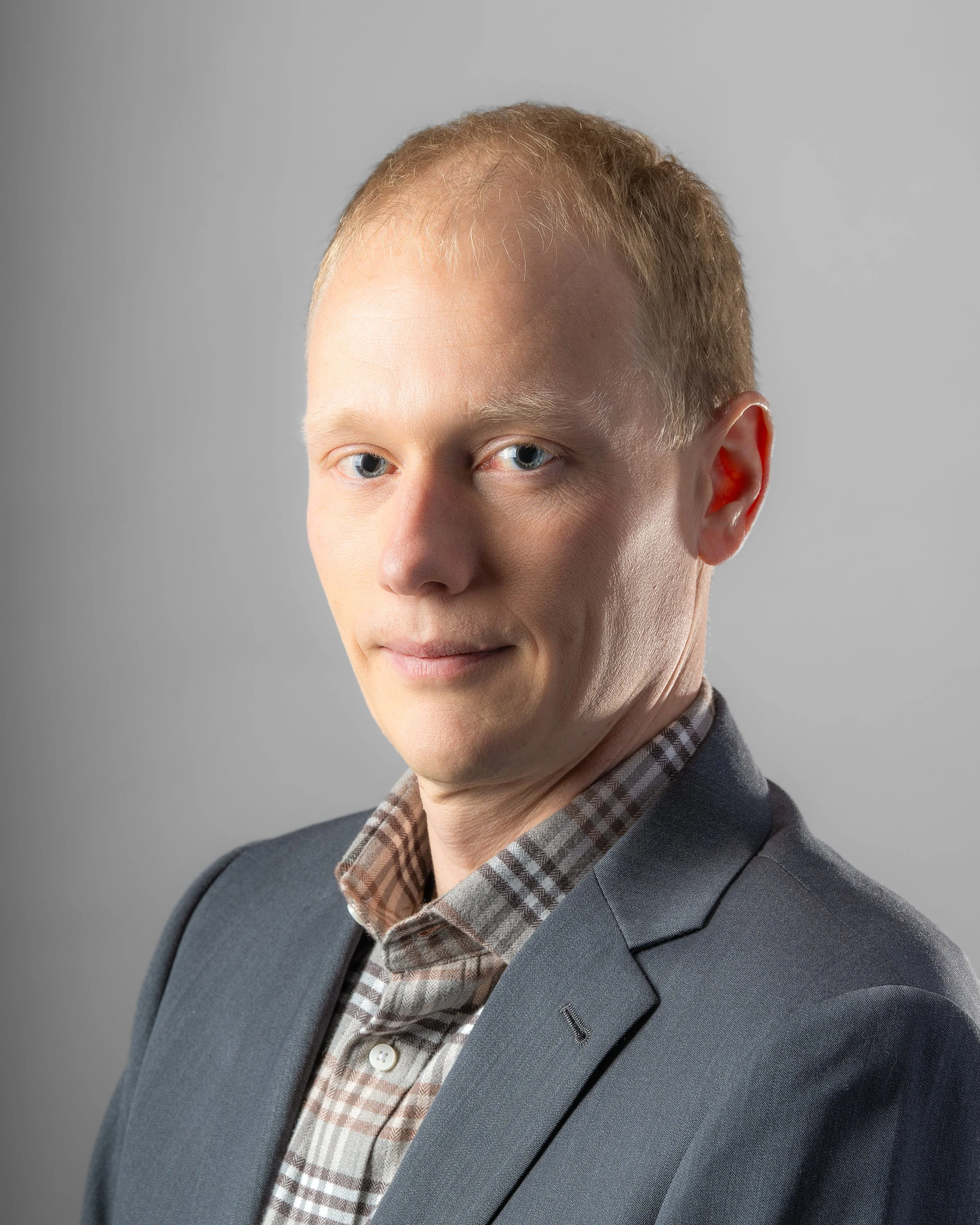 Portrait of a man with light blond hair and blue eyes, wearing a gray blazer and a checkered shirt, against a gray background.