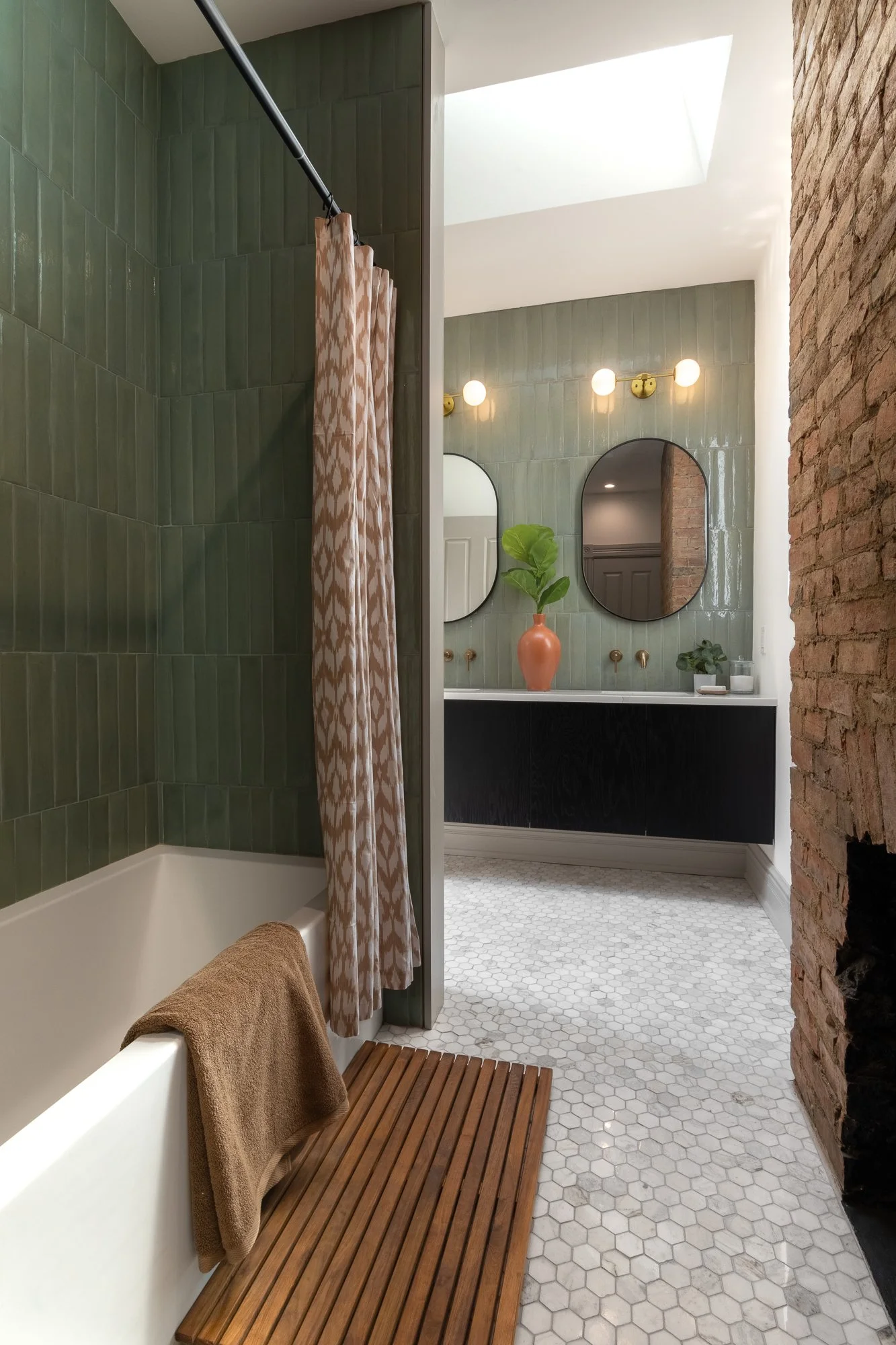 Bathroom with green tiled shower, bathtub with brown towel, wooden bath mat, vanity with black cabinet, round mirrors, plants, and vintage lighting.