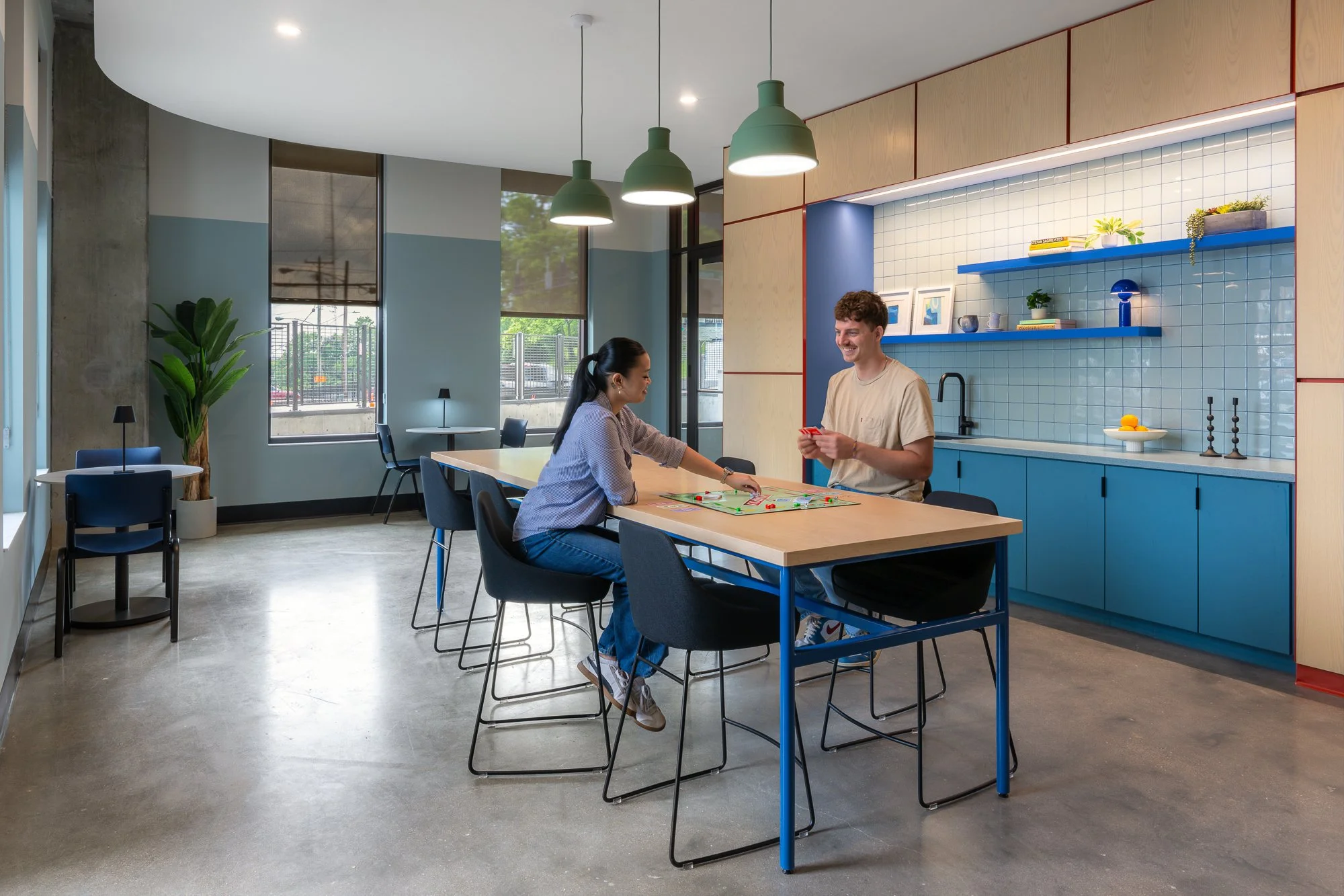 Two people playing a board game at a high table in a modern kitchen or community room with blue and wooden cabinets, blue shelves, and large windows.