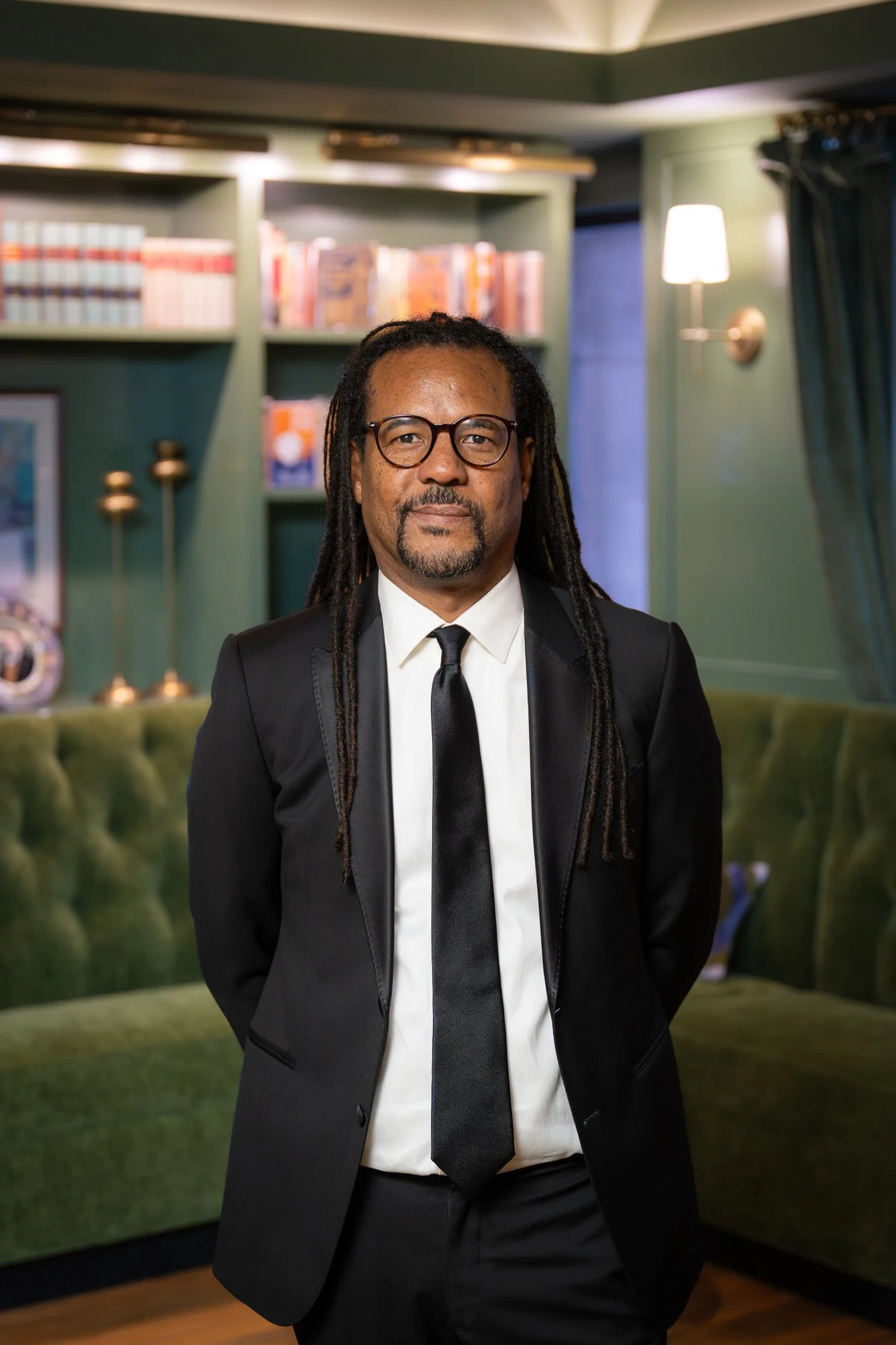 A man with dreadlocks, glasses, and a goatee, dressed in a black suit, white shirt, and black tie, standing in a warmly lit room with green walls, a green velvet sofa, and bookshelves.