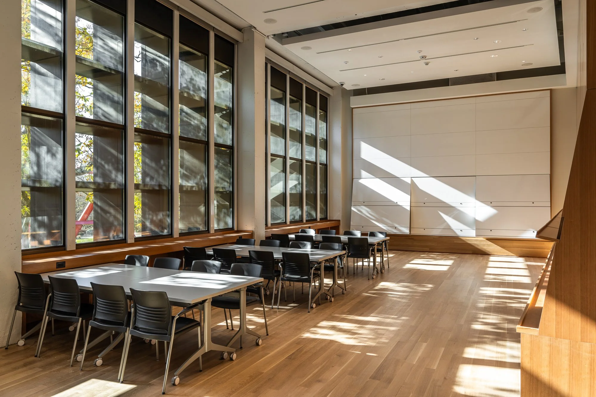 Empty conference or classroom with large windows letting in sunlight, wooden floor, and black chairs around tables.