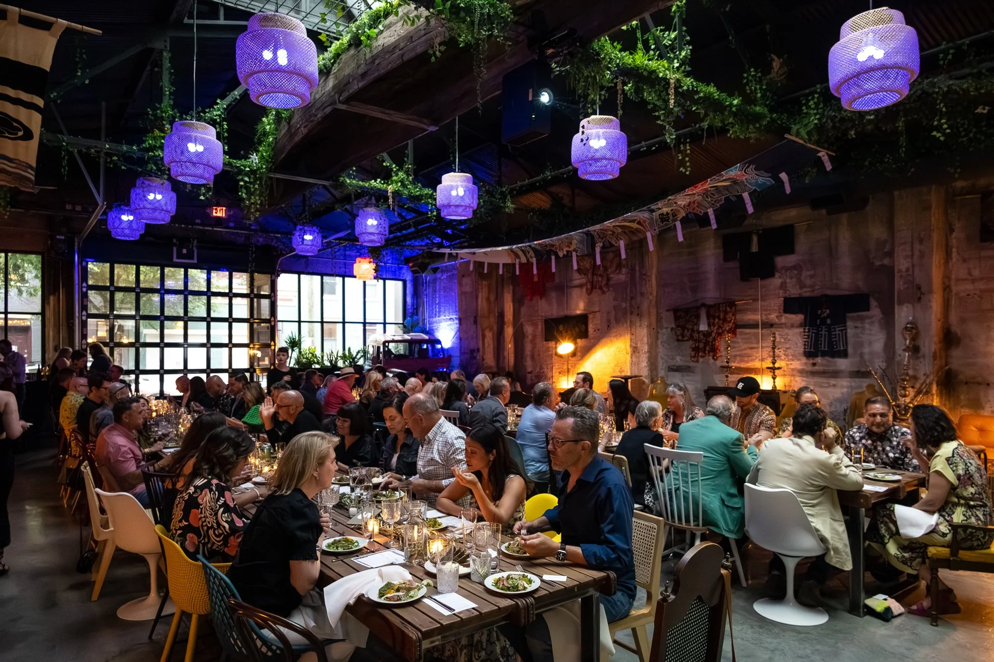 A lively indoor restaurant with a high ceiling, decorated with hanging purple lanterns and greenery. Many people are seated at long tables enjoying a meal, with drinks and plates of food in front of them. The atmosphere is warm and festive, with ambi