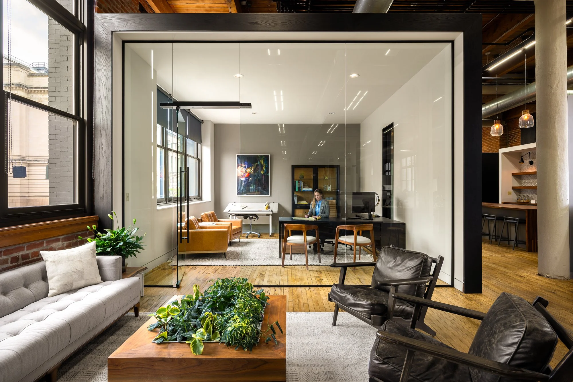 Modern office with glass-walled conference room, natural light, plants, sofa, chairs, and a person working at a desk.