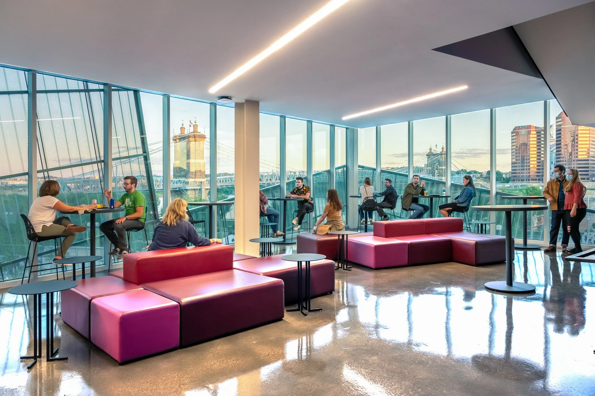 A modern indoor lounge area with pink seating, small round tables, and floor-to-ceiling windows. People are sitting and chatting, some wearing masks. The city skyline and bridge are visible outside.