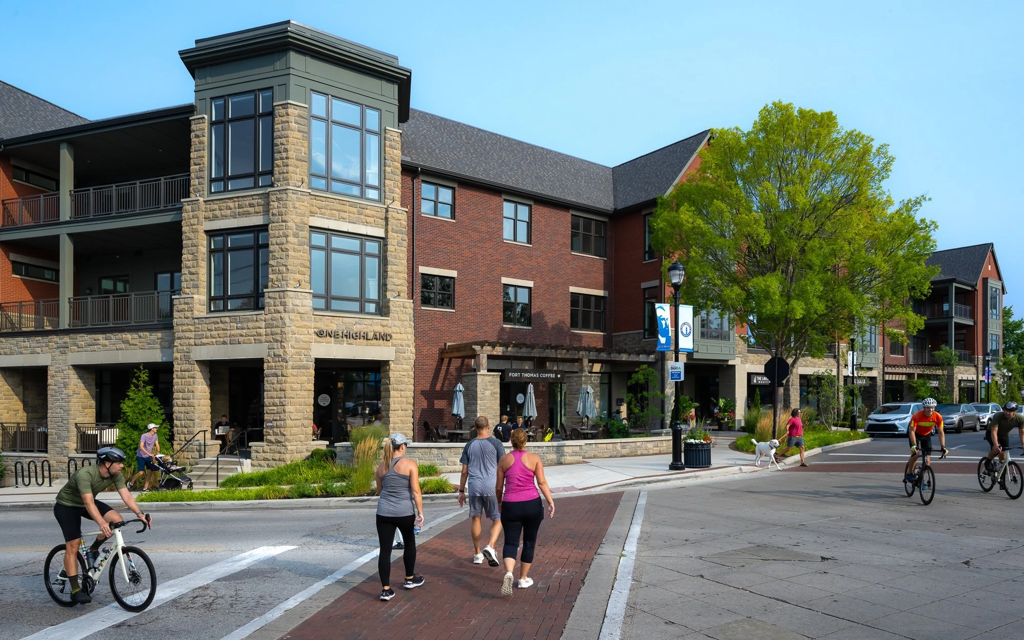 People walking and cycling on a city street in front of a mixed-use building with retail shops and residential units, under a clear blue sky.