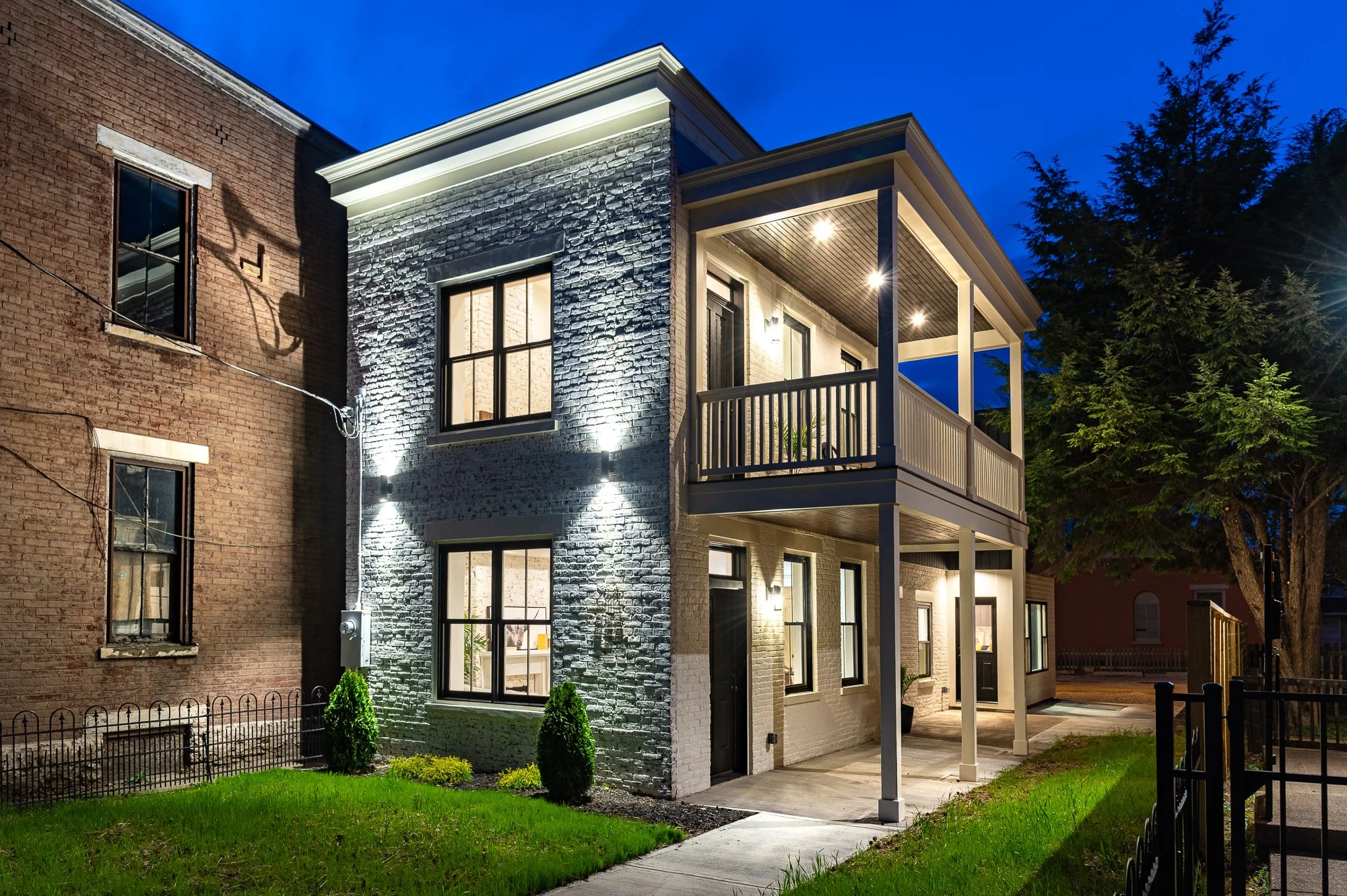A two-story modern house with brick and white painted exterior, illuminated by exterior lights at dusk, featuring a covered balcony on the second floor, a small lawn with bushes, and neighboring buildings on either side.