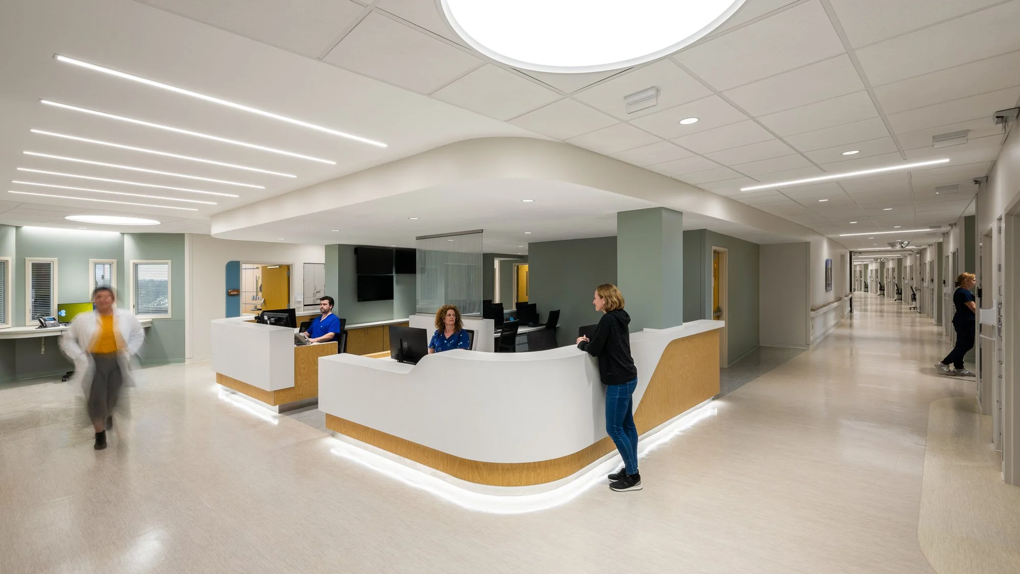 Hospital reception area with a curved white front desk, healthcare staff working at computers, and a hallway with patient rooms to the right.