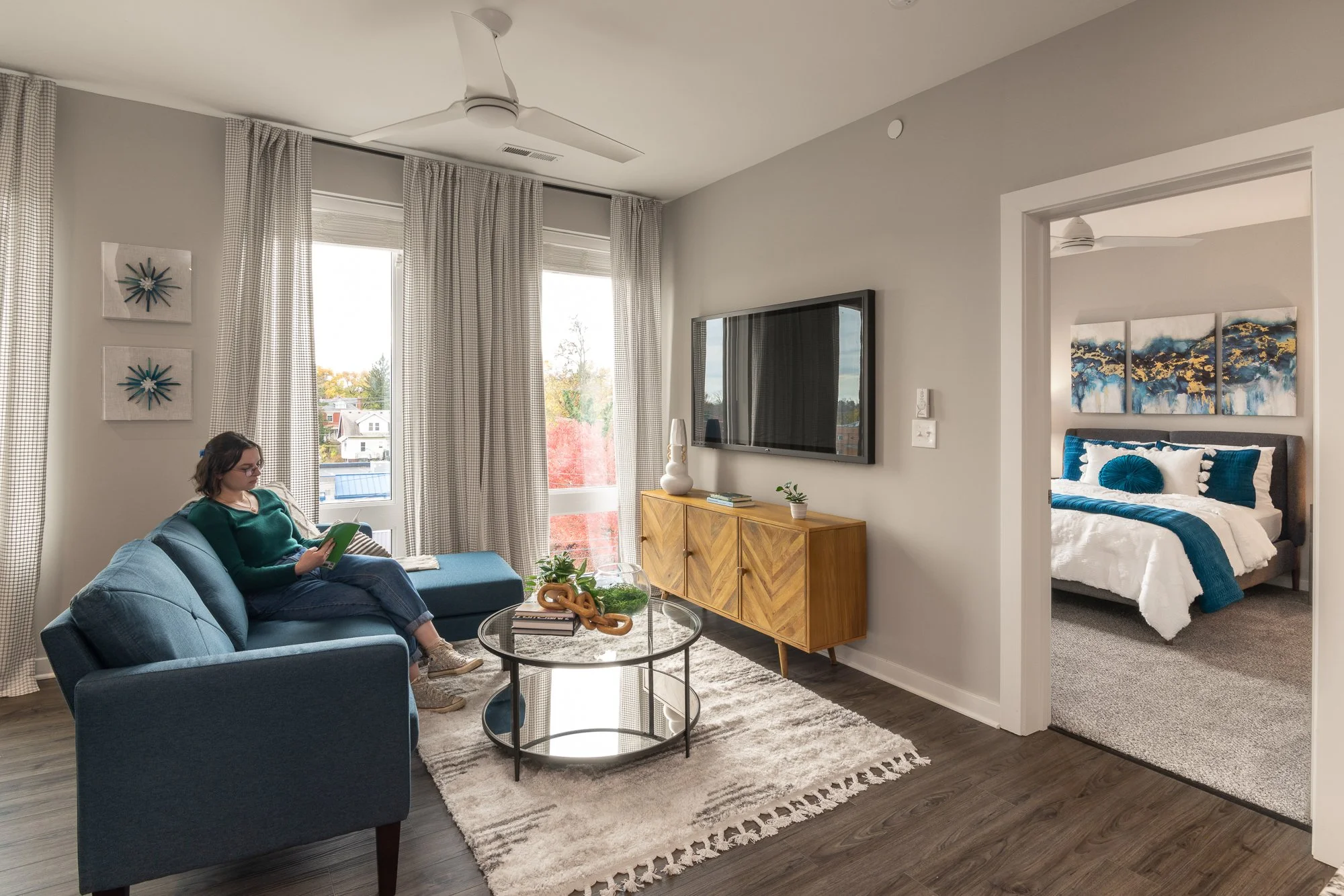 Living room with blue sofa, woman reading a book, large windows with curtains, wall art, coffee table with decorations, wooden TV stand, flat-screen TV, and a bedroom visible through a doorway featuring a bed with blue bedding and wall art.