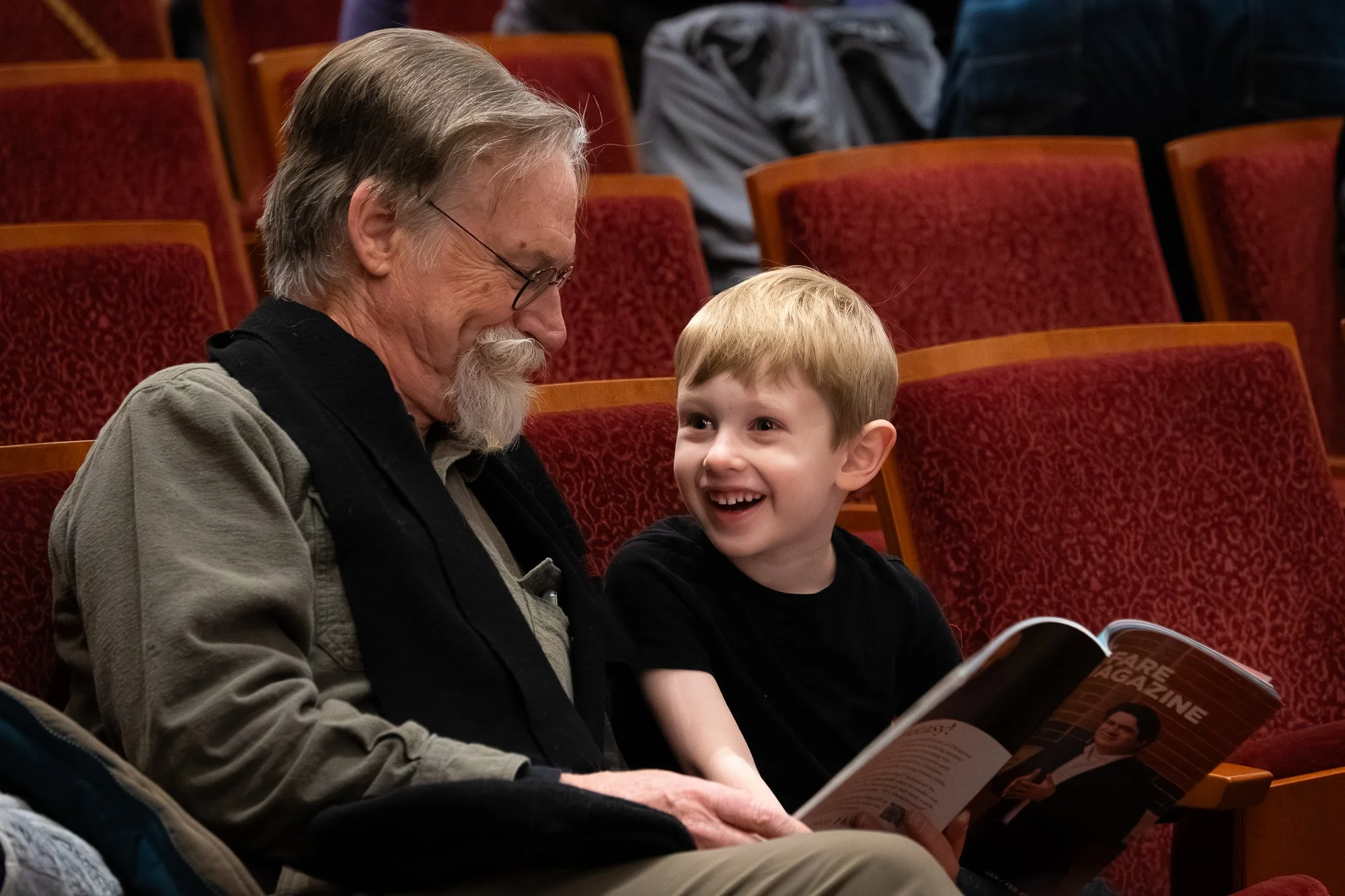 An elderly man and a young boy sitting together in an auditorium, smiling and sharing a magazine.