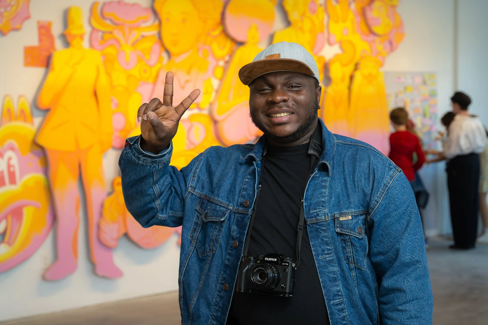 A man smiling and making a peace sign with his right hand at an art gallery. He is wearing a denim jacket, a black shirt, a camera around his neck, and a cap. Behind him are colorful abstract paintings and people viewing art.