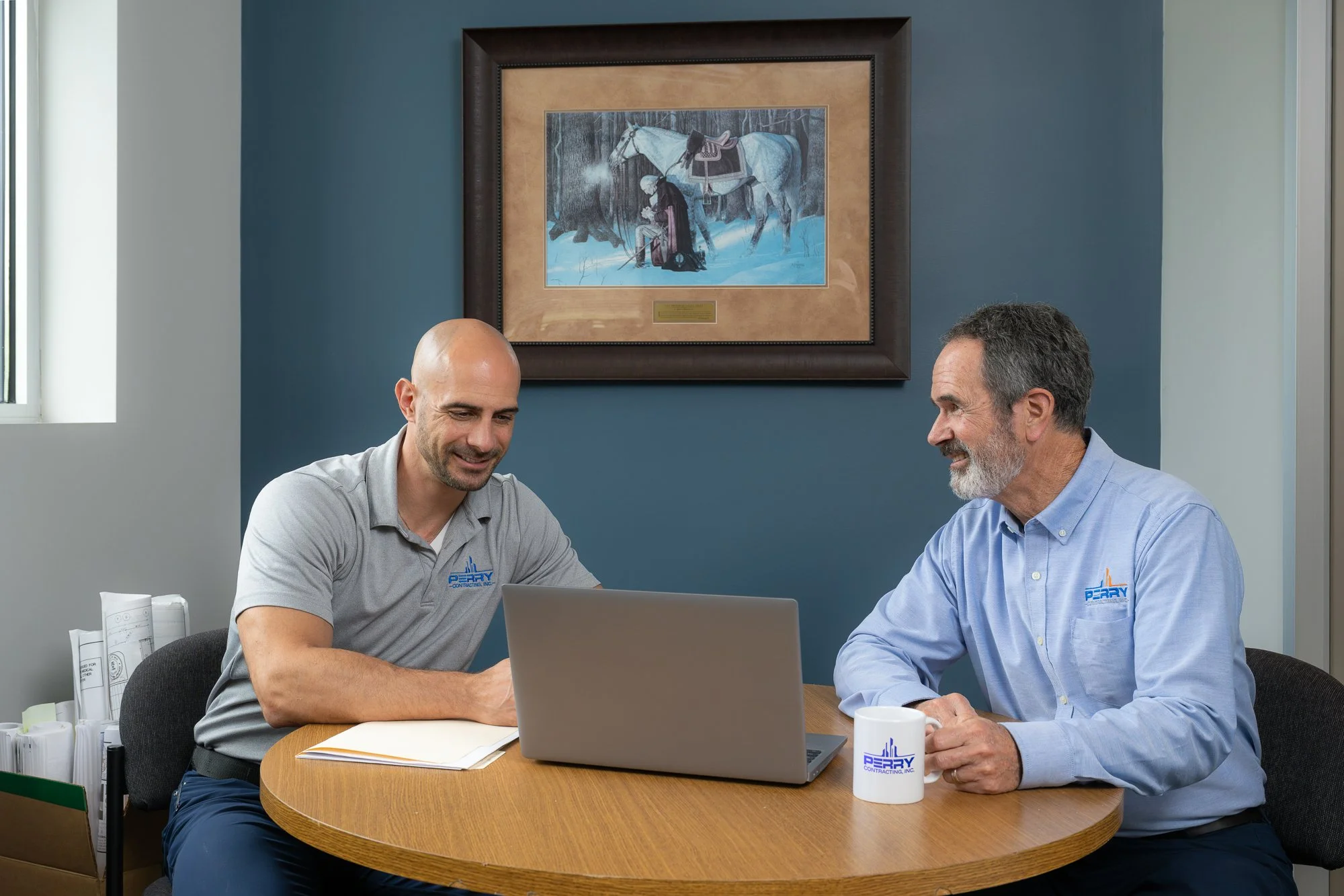 Two men sitting at a round wooden table in an office, looking at a laptop screen and smiling. One man has a bald head, the other has gray hair and a beard. Both are wearing shirts with a company logo. There is artwork of a person and a horse hanging 