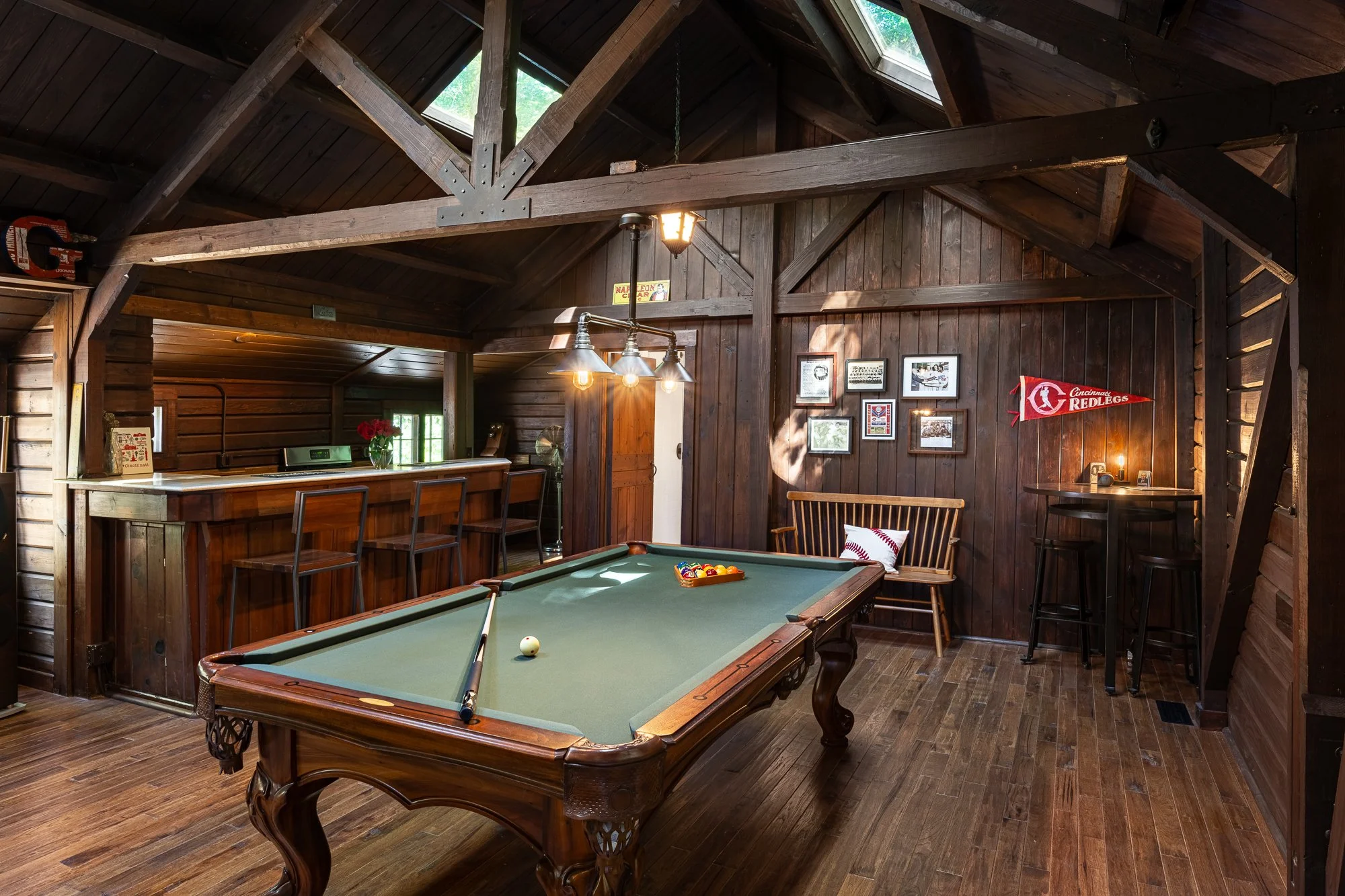Impressive wooden game room featuring a classic pool table, bar counter with three chairs, wall decorated with framed pictures and a Cincinnati Reds pennant, and a wooden bench with pillow. The room has a vaulted ceiling with skylights and is illumin