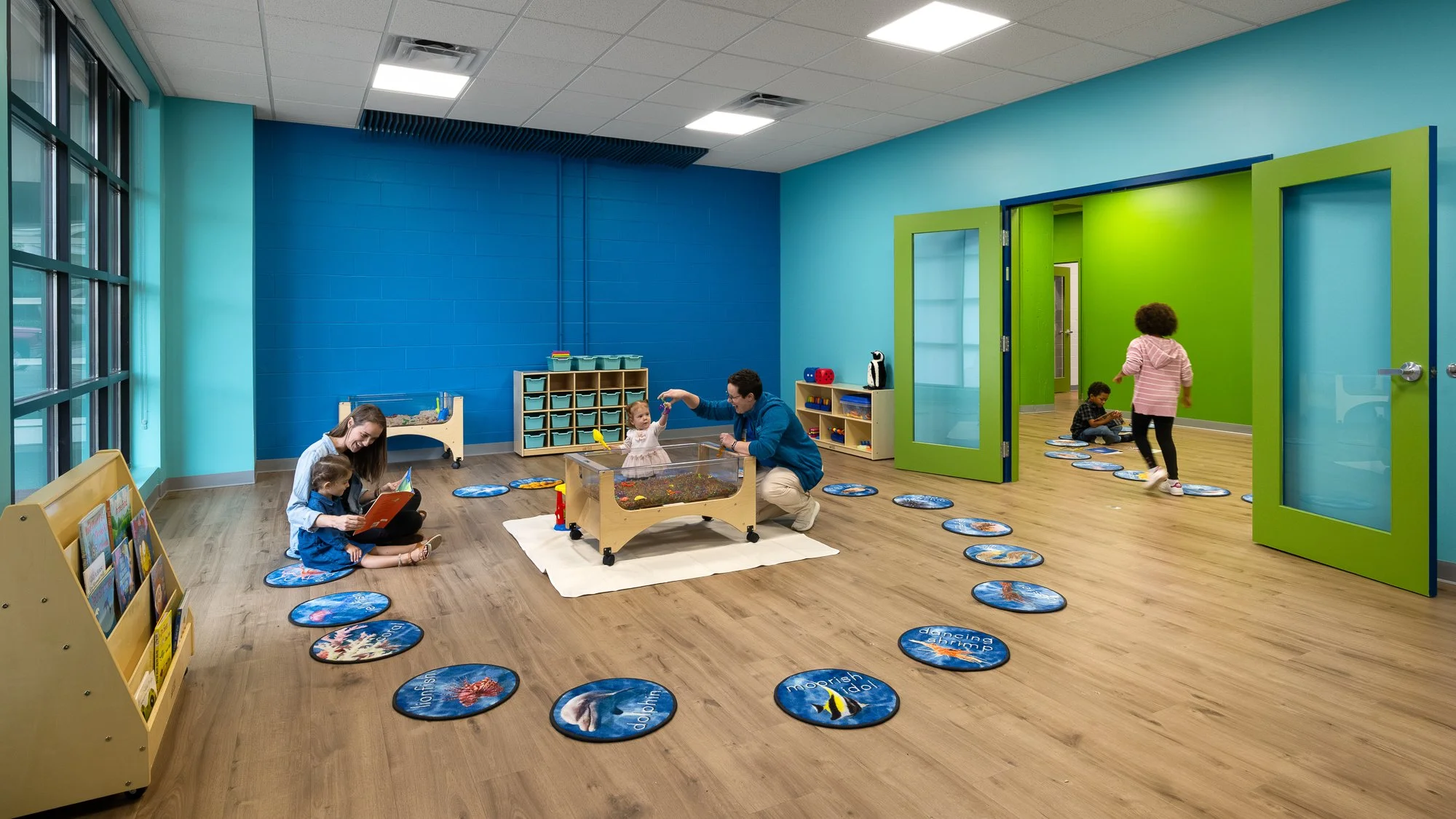 Children and a caregiver in a colorful indoor playroom with wooden flooring, blue and green walls, large windows, and various toys, books, and a feeding tub for aquatic animals.