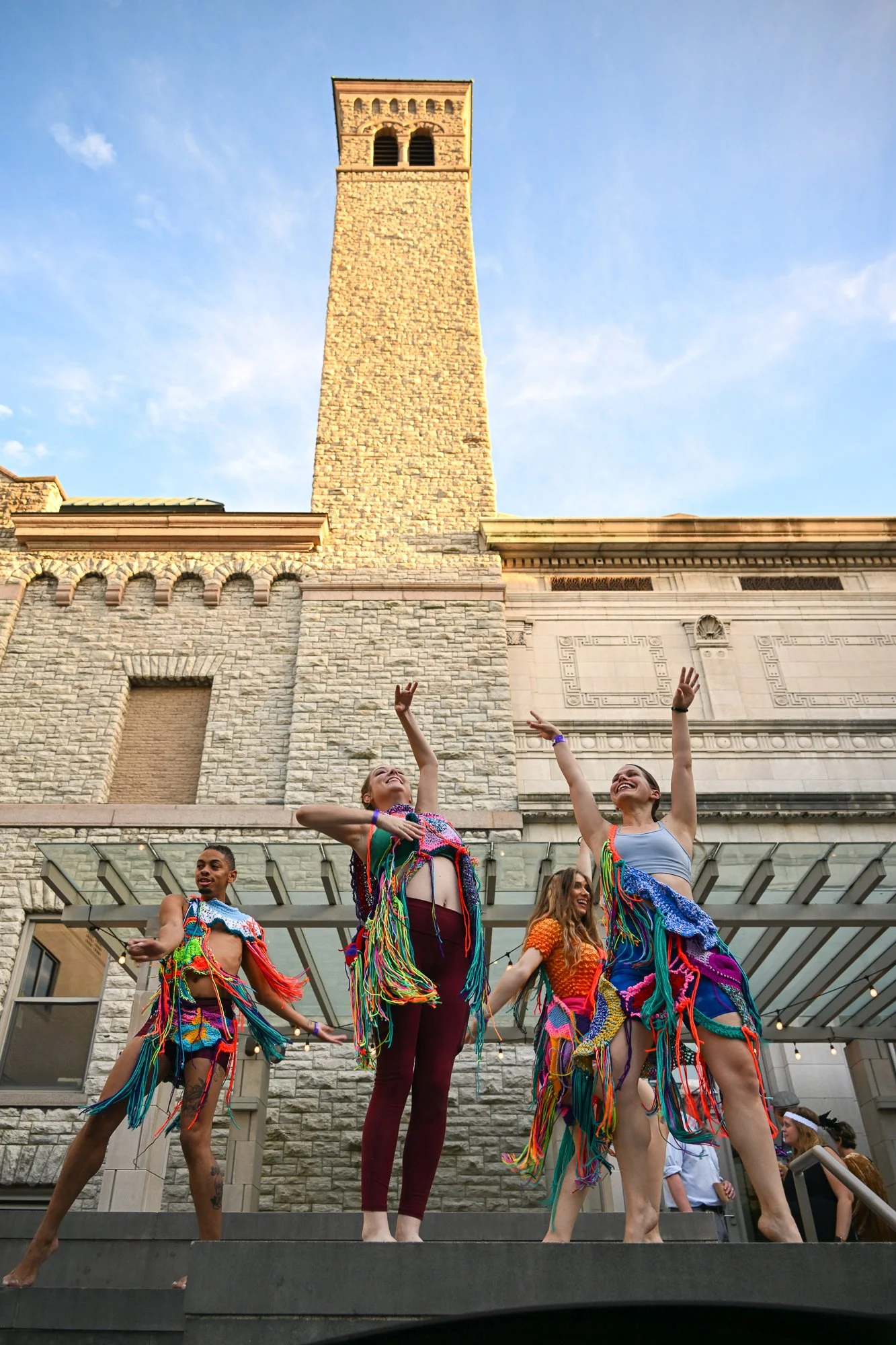 Four dancers wearing colorful, fringed costumes perform outdoors in front of a stone building with a tall bell tower. They are smiling and striking dance poses against a blue sky.