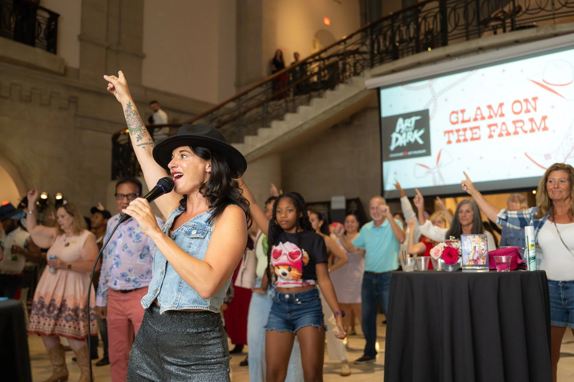 A woman with dark hair, wearing a black hat, denim vest, and sparkly skirt, singing into a microphone at a lively indoor event with a diverse group of people dancing and raising their hands in the background, with a large screen reading 'Glam on the 