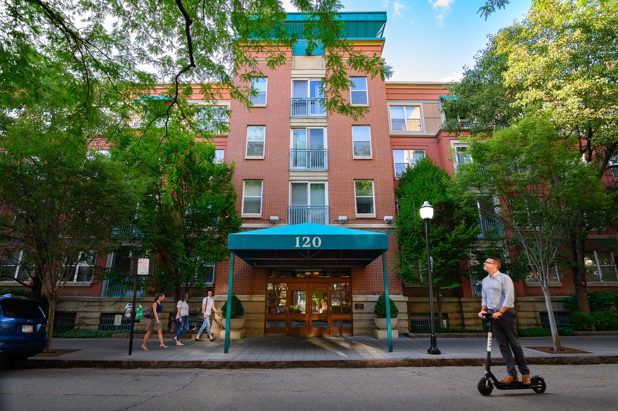 Multi-story brick apartment building with a blue awning displaying the number 120, surrounded by green trees, with pedestrians walking on the sidewalk and a man riding an electric scooter in front.